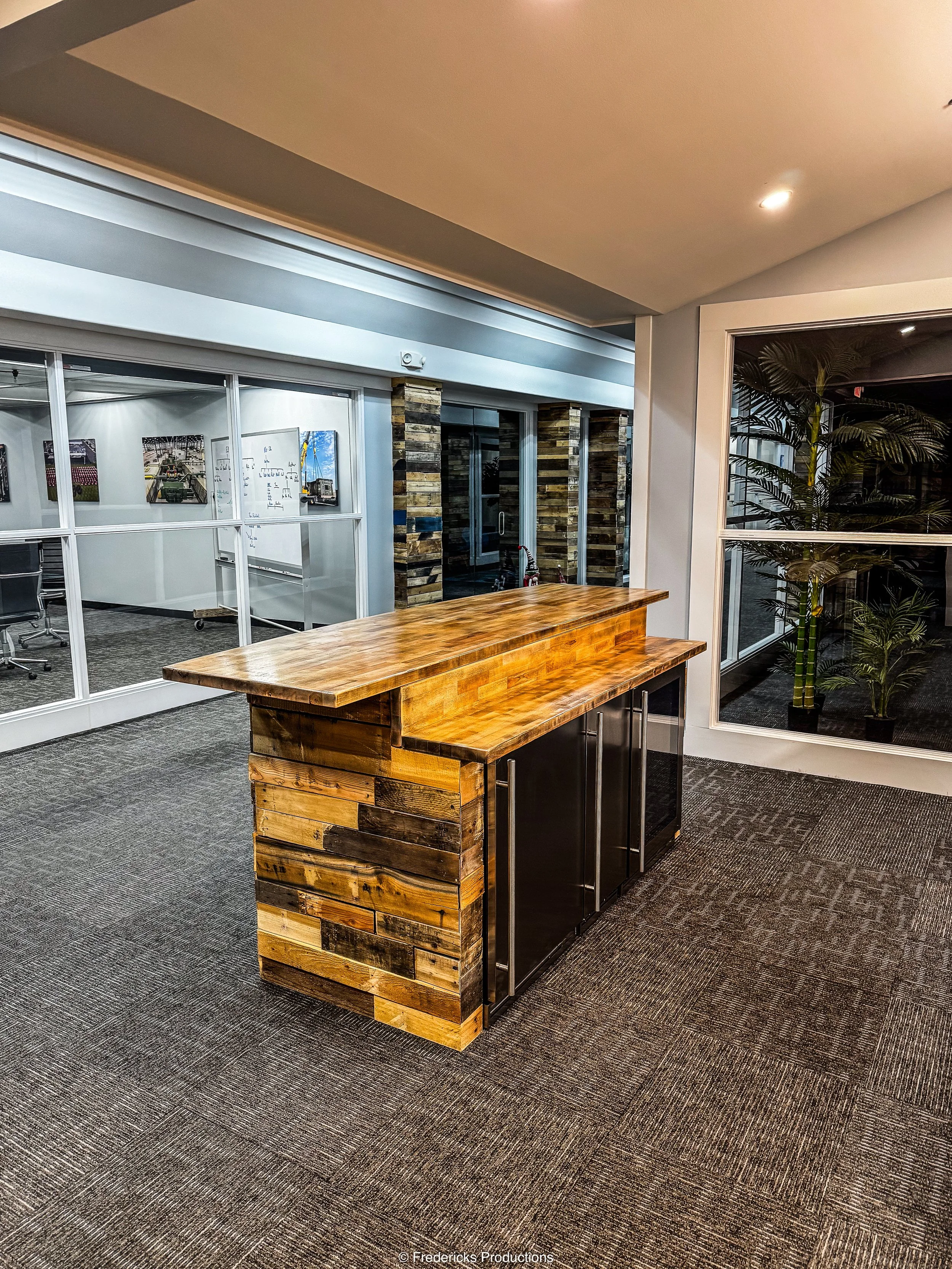 Modern office reception area with a wooden countertop and cabinets, glass partition walls, potted plants, and carpeting.