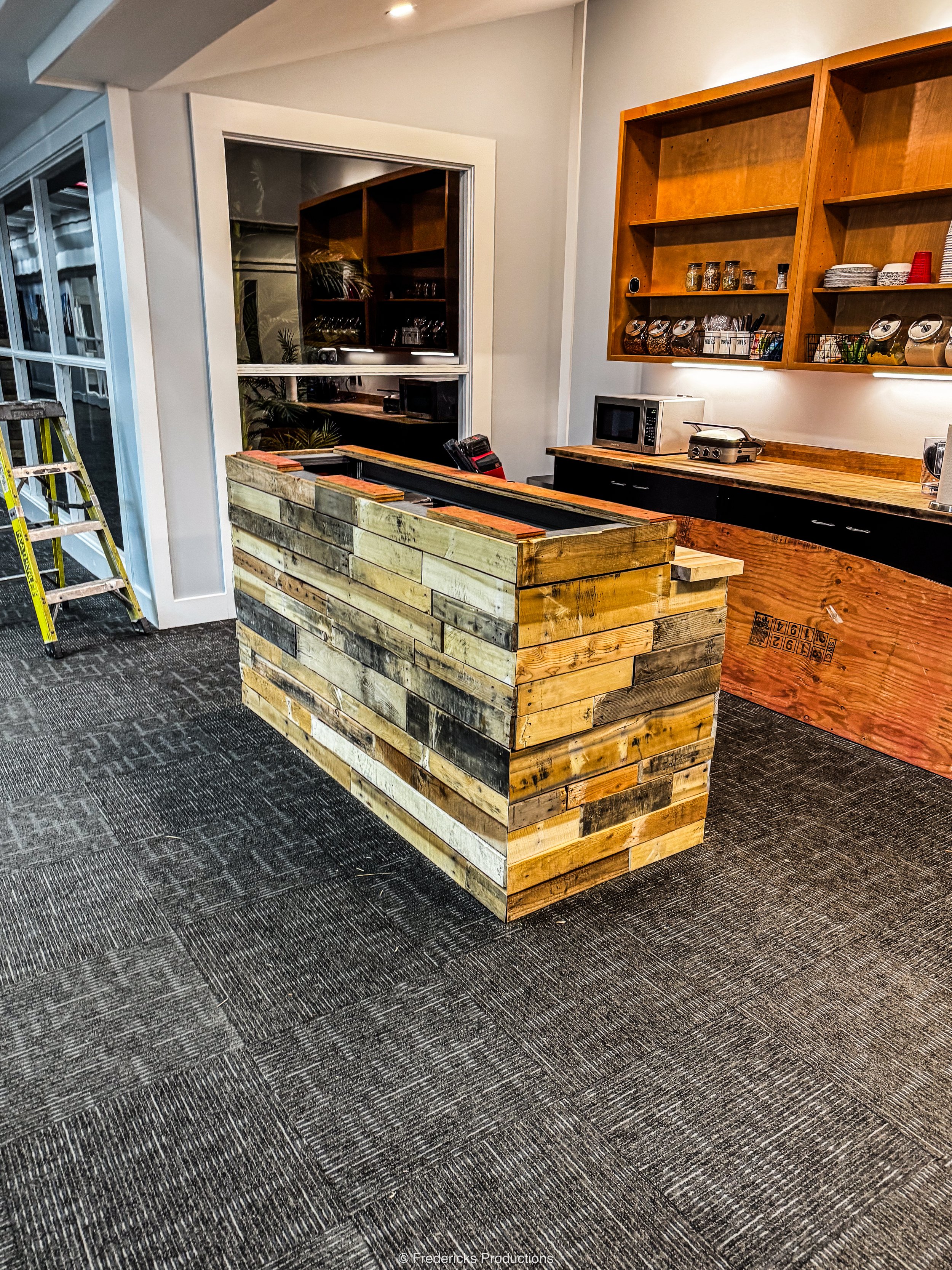 Interior of a coffee shop or cafe with a wooden counter made from pallet wood, open shelving on the wall displaying jars, dishes, and cups, a microwave, a small oven, and a ladder leaning against the wall.