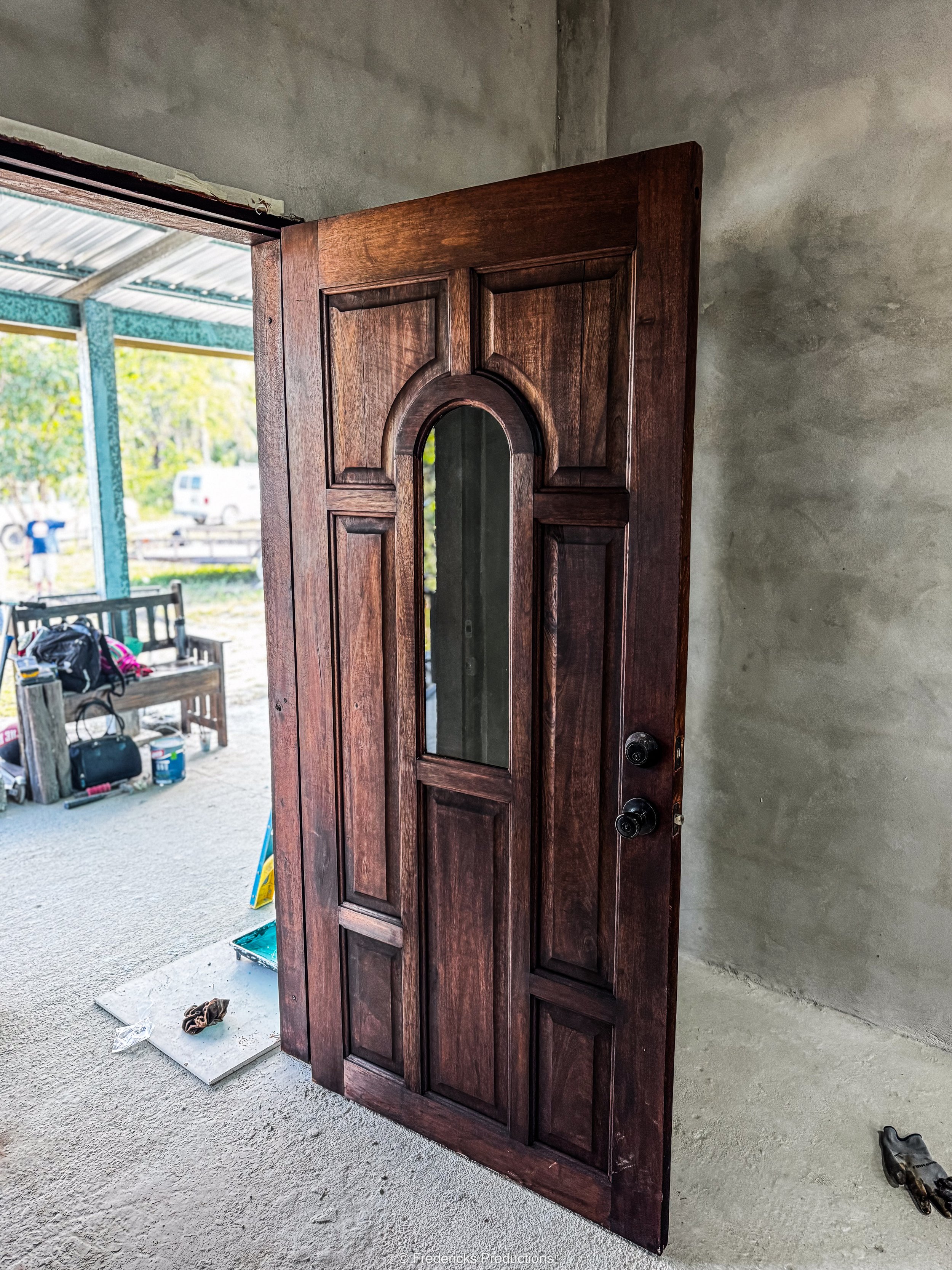 A wooden front door with an arched window panel leaning against a concrete wall, partially open, revealing an outdoor bench and a view of trees and vehicles.