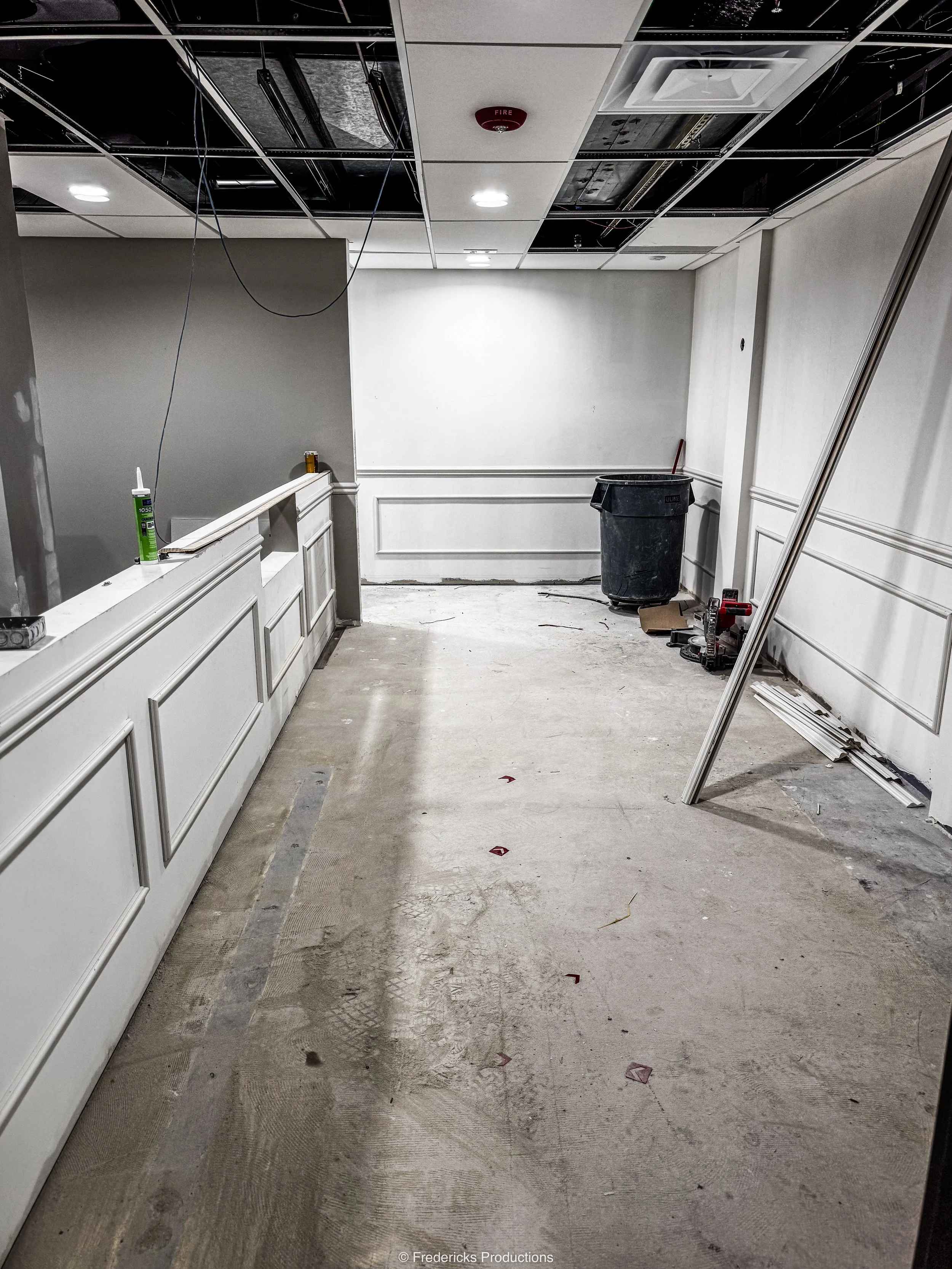 Interior of a building under construction or renovation with unfinished ceiling, disorganized workspace, cleaning supplies, and construction tools on the floor.