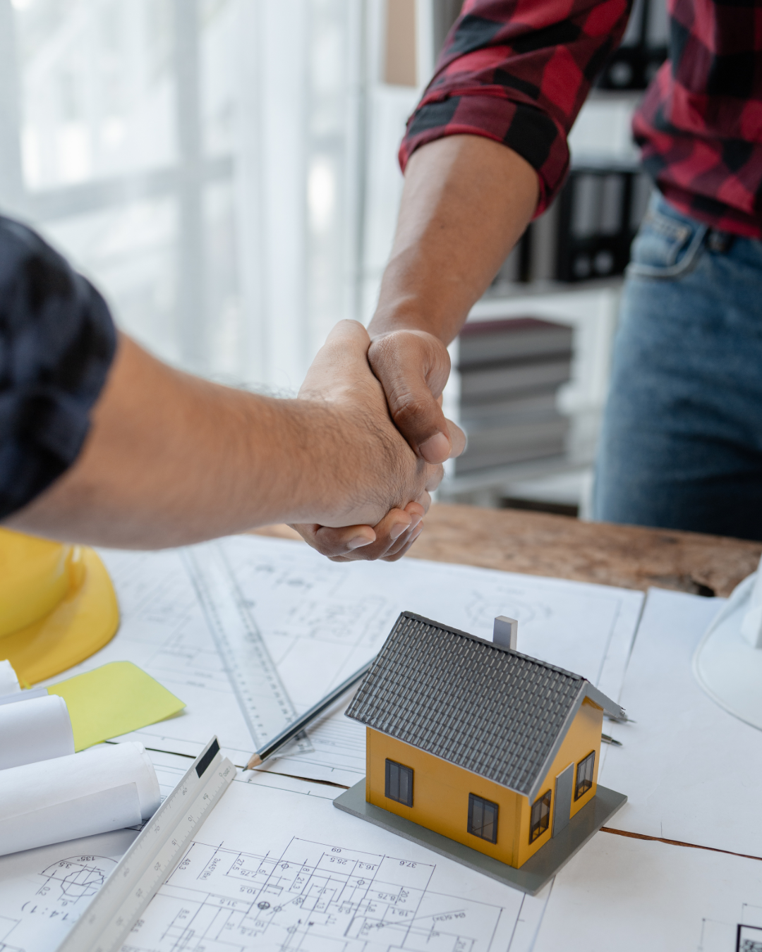 Two people shaking hands over architectural plans and a small model house on a worktable.