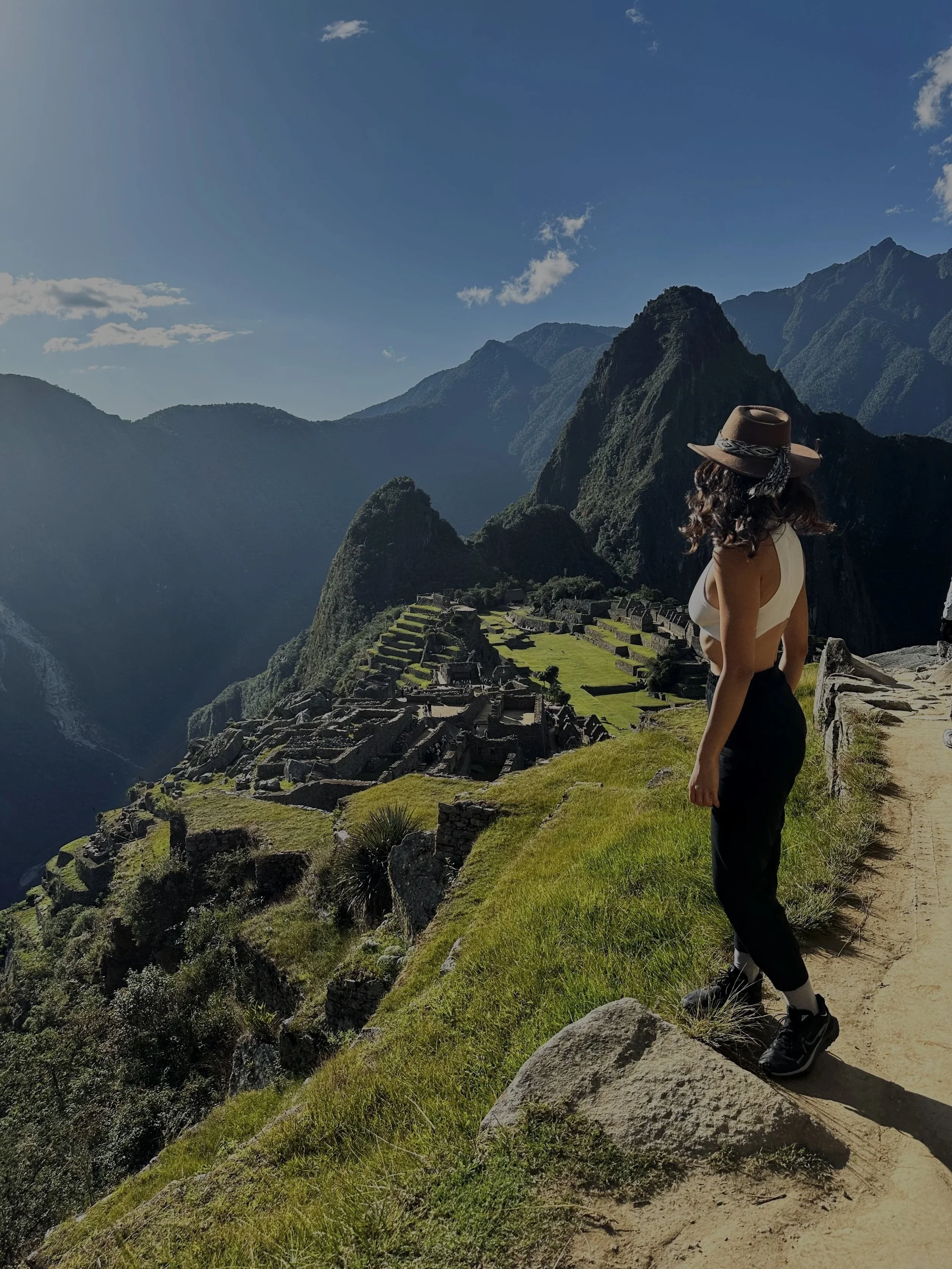 A woman wearing a brown hat, white top, black pants, and sneakers standing on a trail overlooking Machu Picchu with mountains in the background.