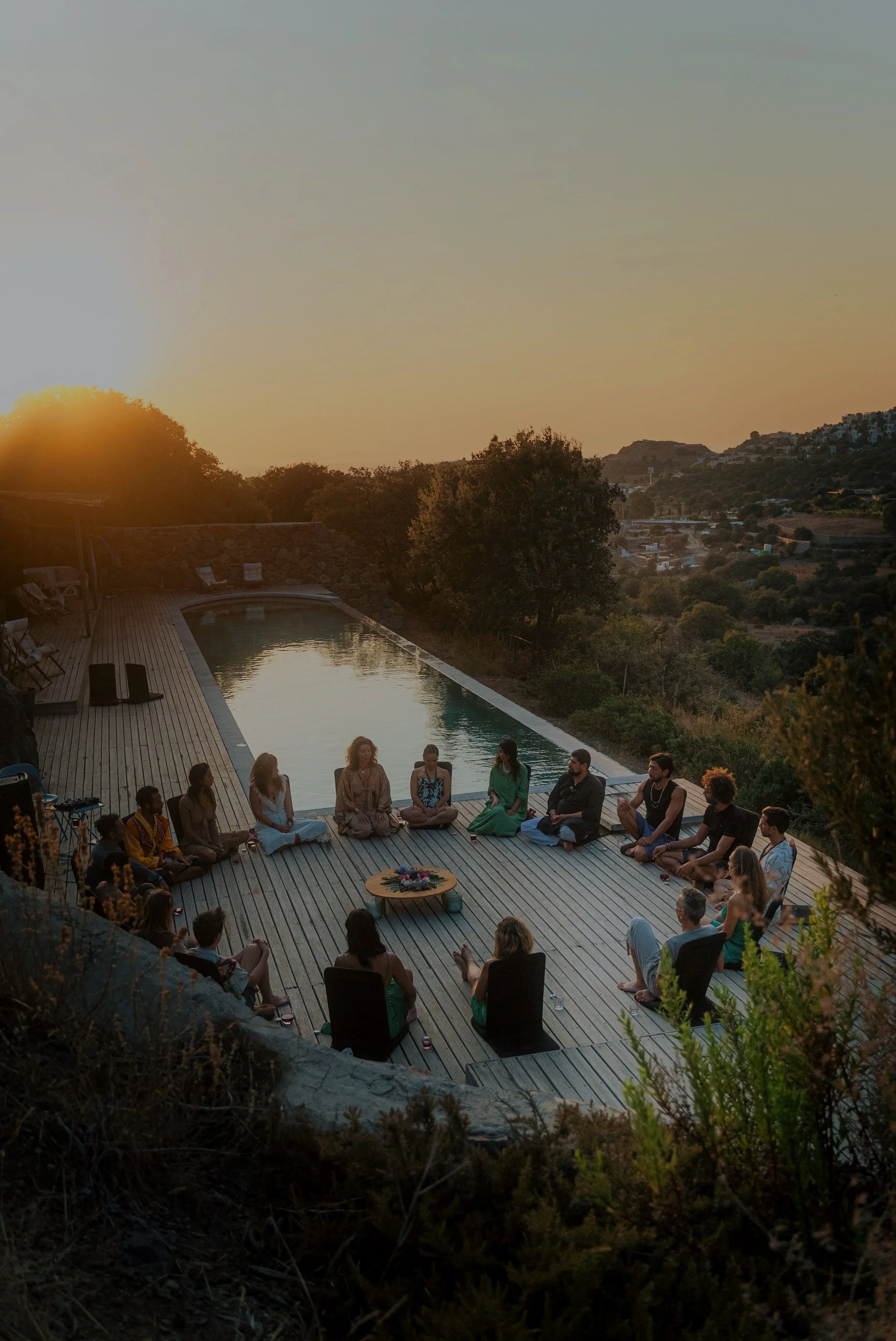 People sitting in a circle on a wooden deck near a swimming pool at sunset.