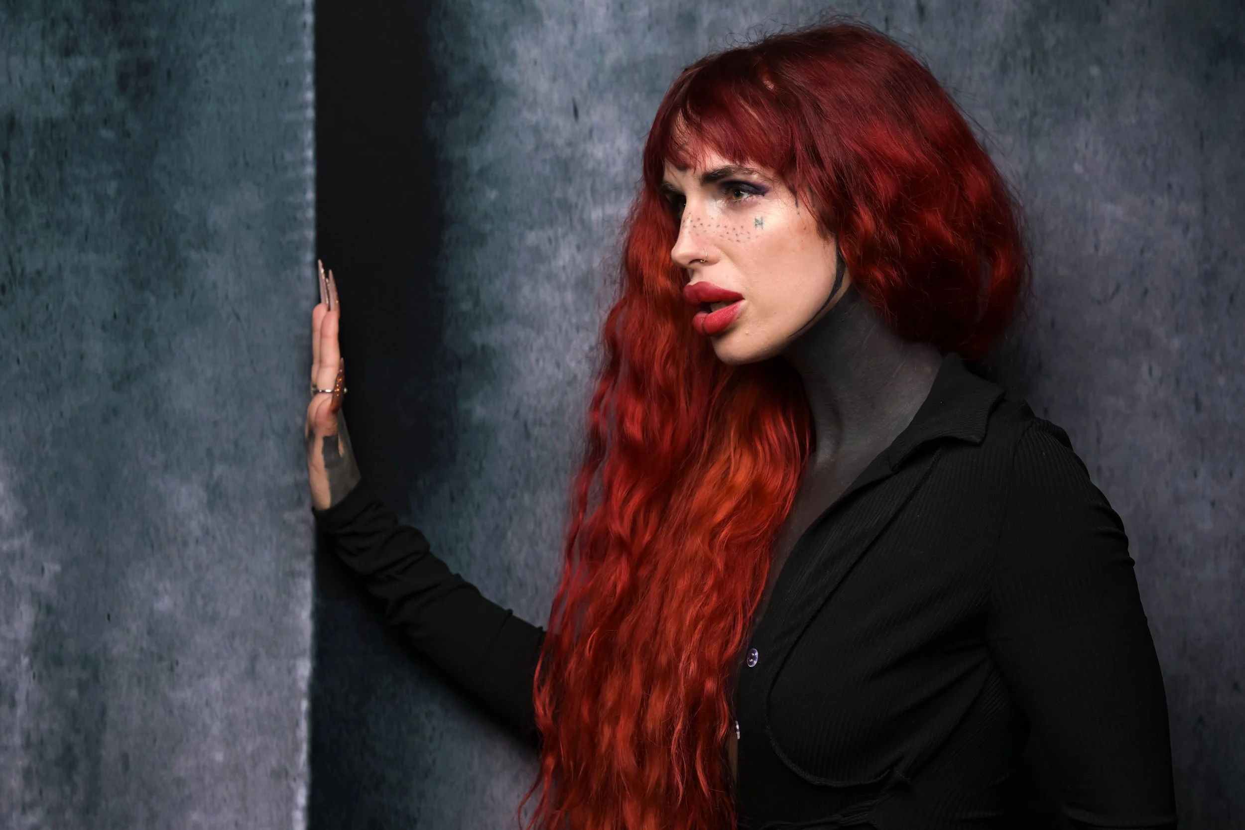 A woman with long, wavy red hair, makeup with freckles, and dark lipstick leans against a textured gray wall, wearing a black long-sleeve shirt.