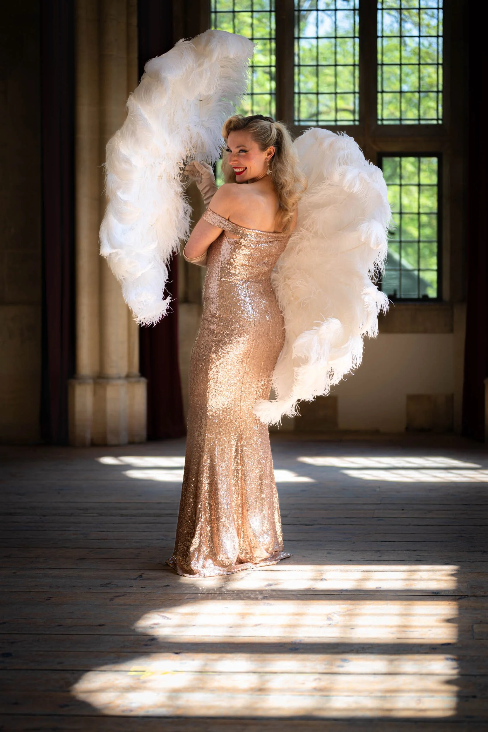 A woman in a shimmering gold gown with large white feathered wings, smiling and posing indoors with sunlight streaming through windows behind her.