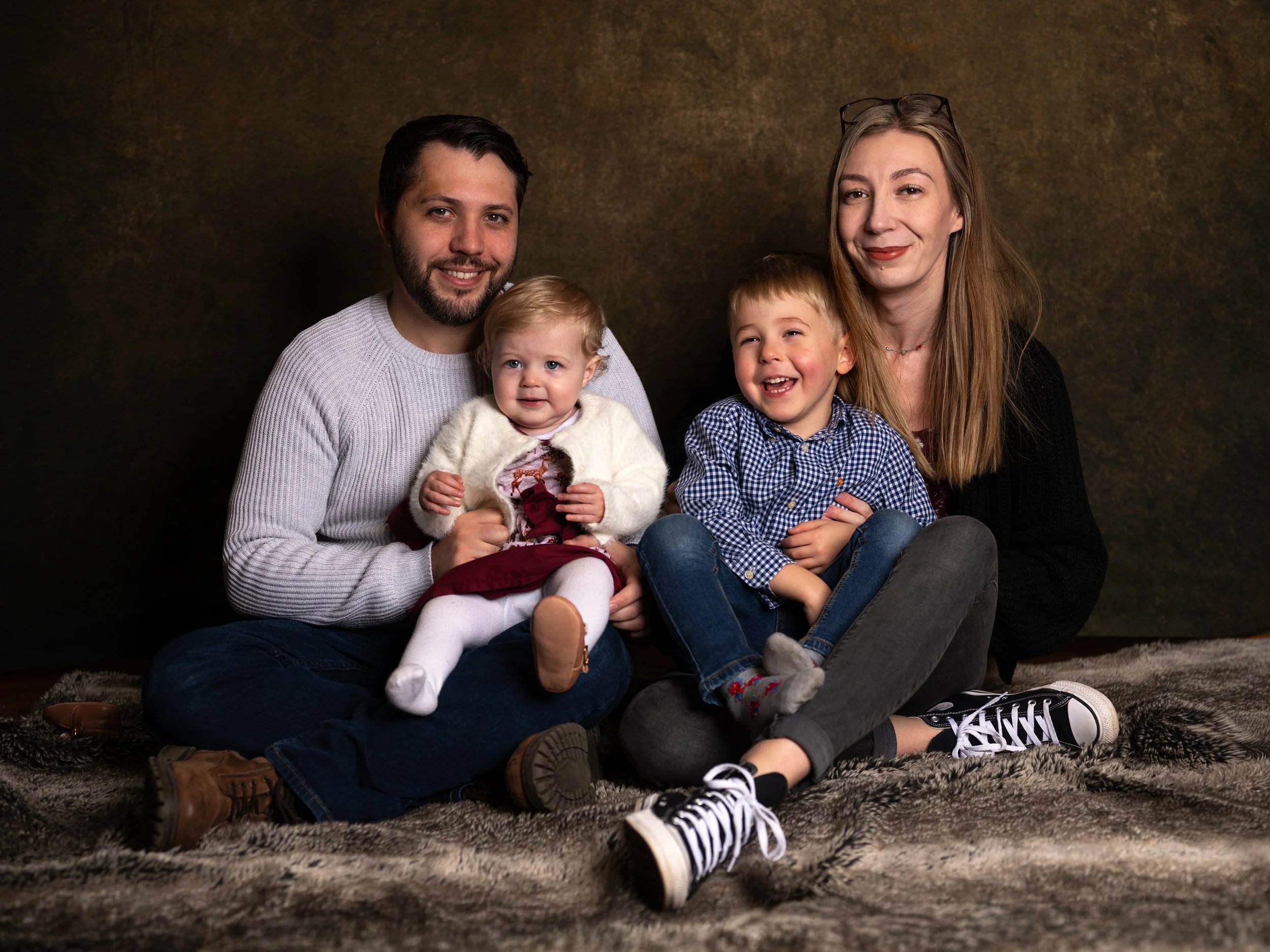 A family of four sitting on a fur rug poses for a photo. The father has dark hair and a beard, wearing a gray sweater, and holds a young girl with blonde hair. The daughter is wearing a white cardigan, maroon dress, and white tights. The mother has light brown hair, glasses on her head, and wears a black jacket. She is sitting next to a young boy with blonde hair, wearing a blue checkered shirt and jeans. The background is dark and textured.