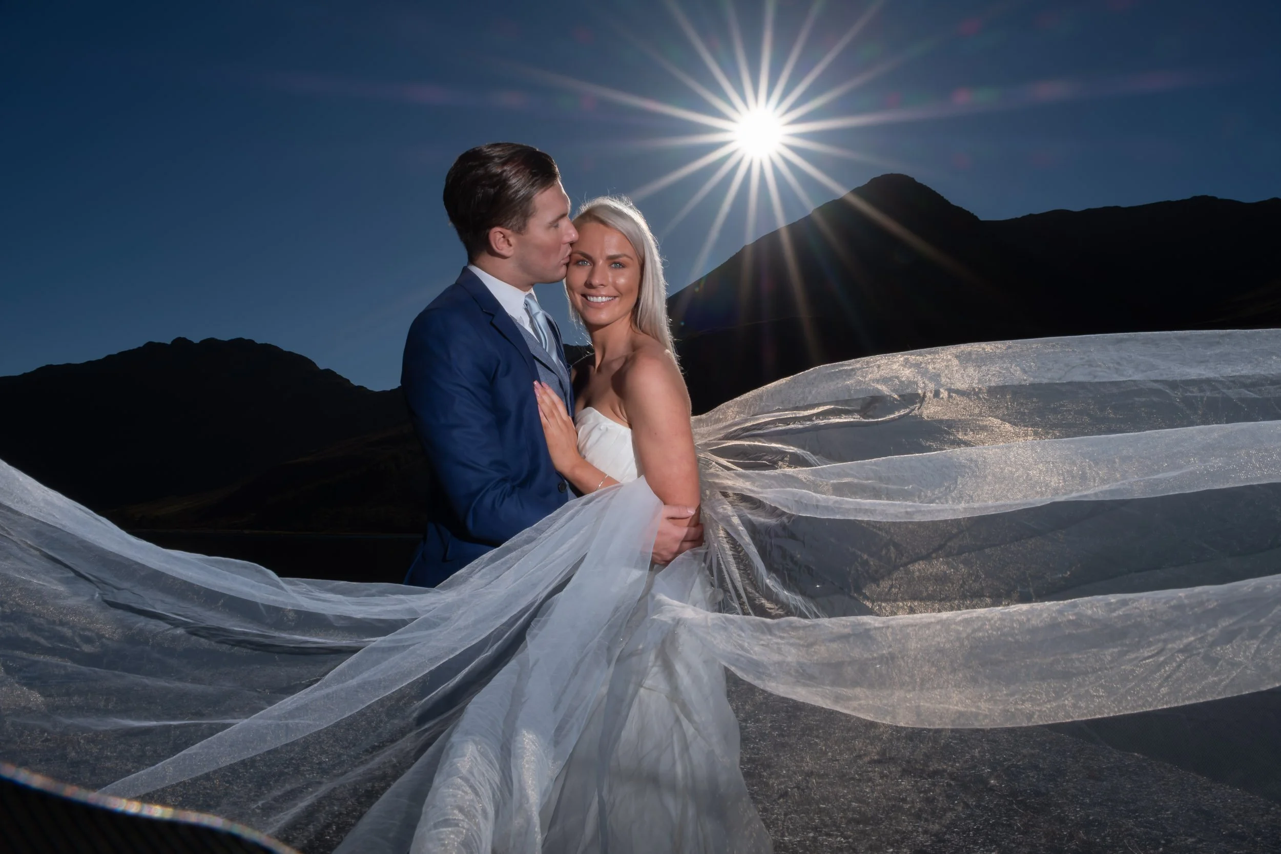 A bride and groom in wedding attire embracing outdoors under a bright sun with mountains in the background.