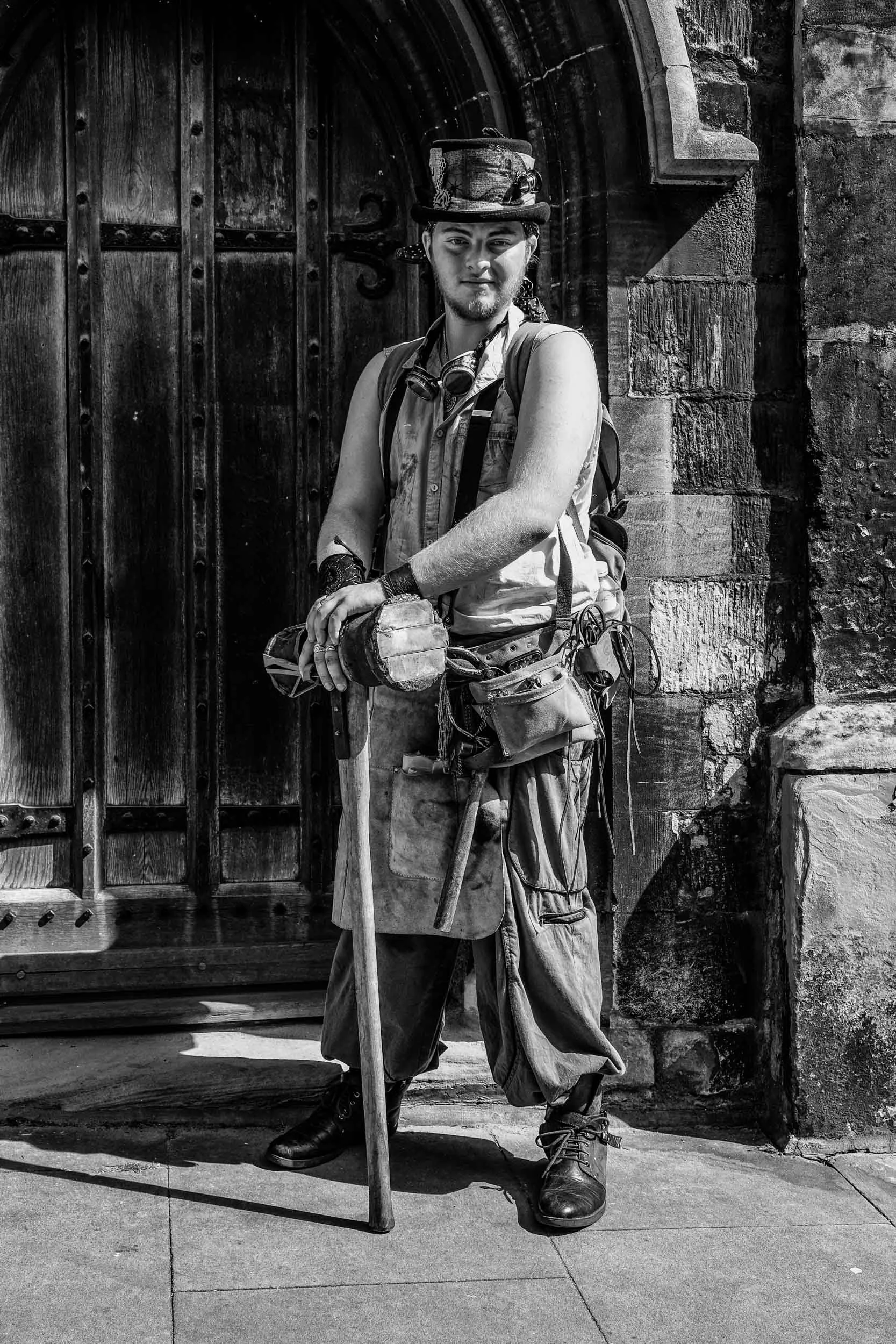 A young person dressed as a steampunk character, wearing a top hat with various accessories, standing in front of a wooden door. They have a confident expression, gloves, goggles around their neck, and are holding a walking stick, dressed in a sleeve