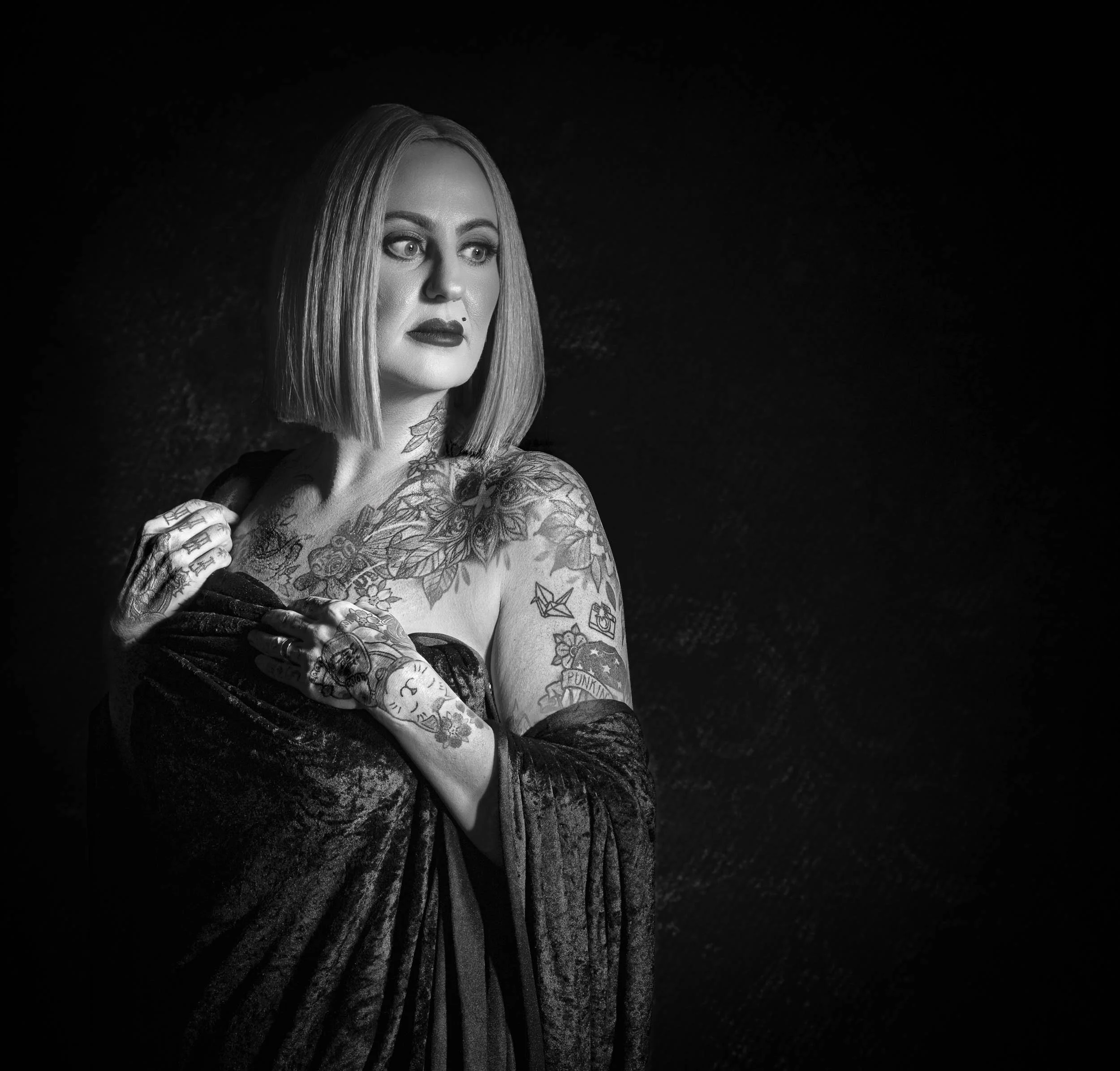 Black and white portrait of a woman with tattoos, short hair, wearing a dark off-the-shoulder dress, looking to the side with a serious expression.