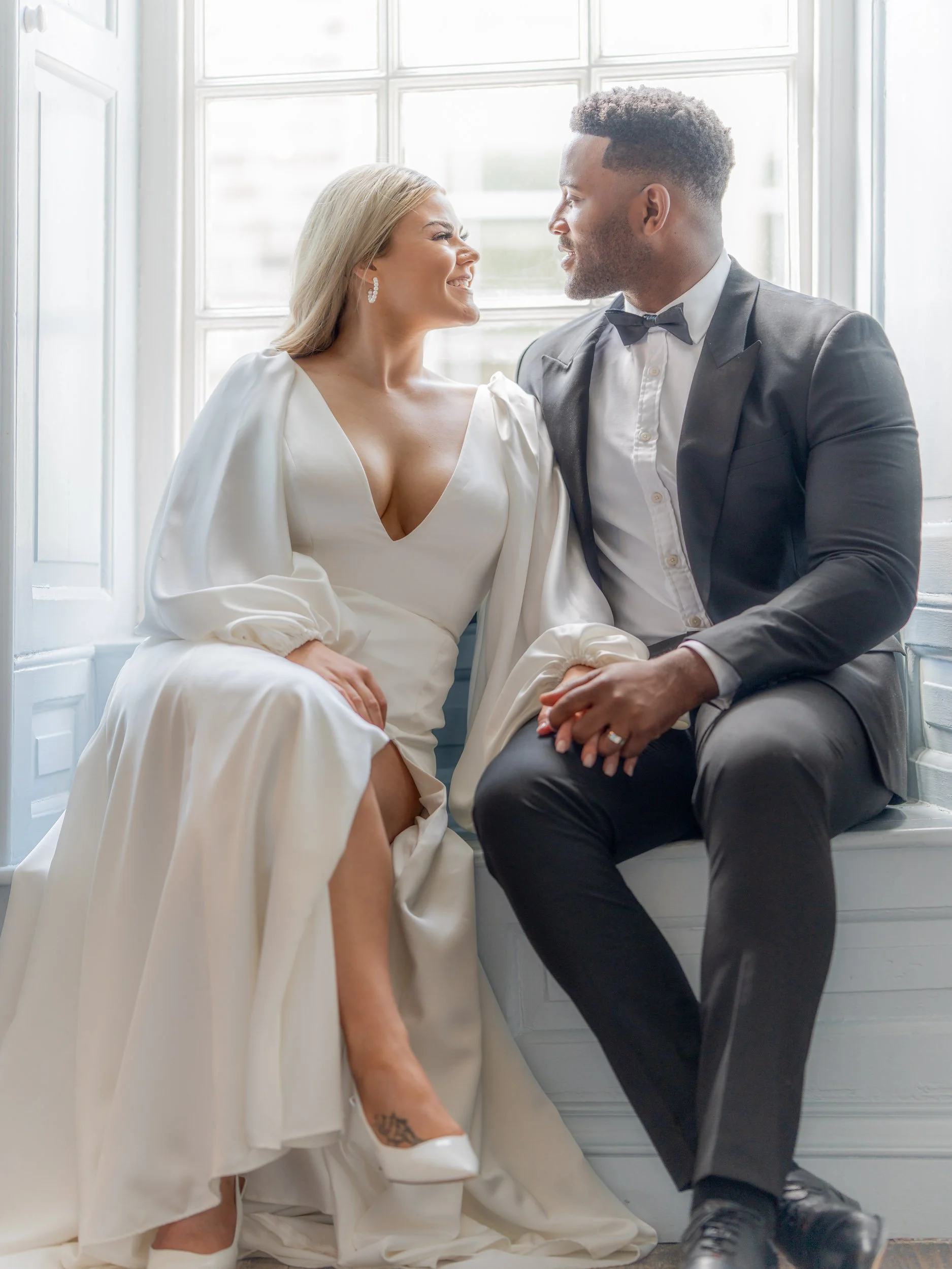 A bride and groom sitting close together near a large window, gazing into each other's eyes. The bride wears a white wedding gown with a deep neckline, and the groom wears a black tuxedo with a bow tie. They are smiling and holding hands, with the bride's high-heeled shoes visible.