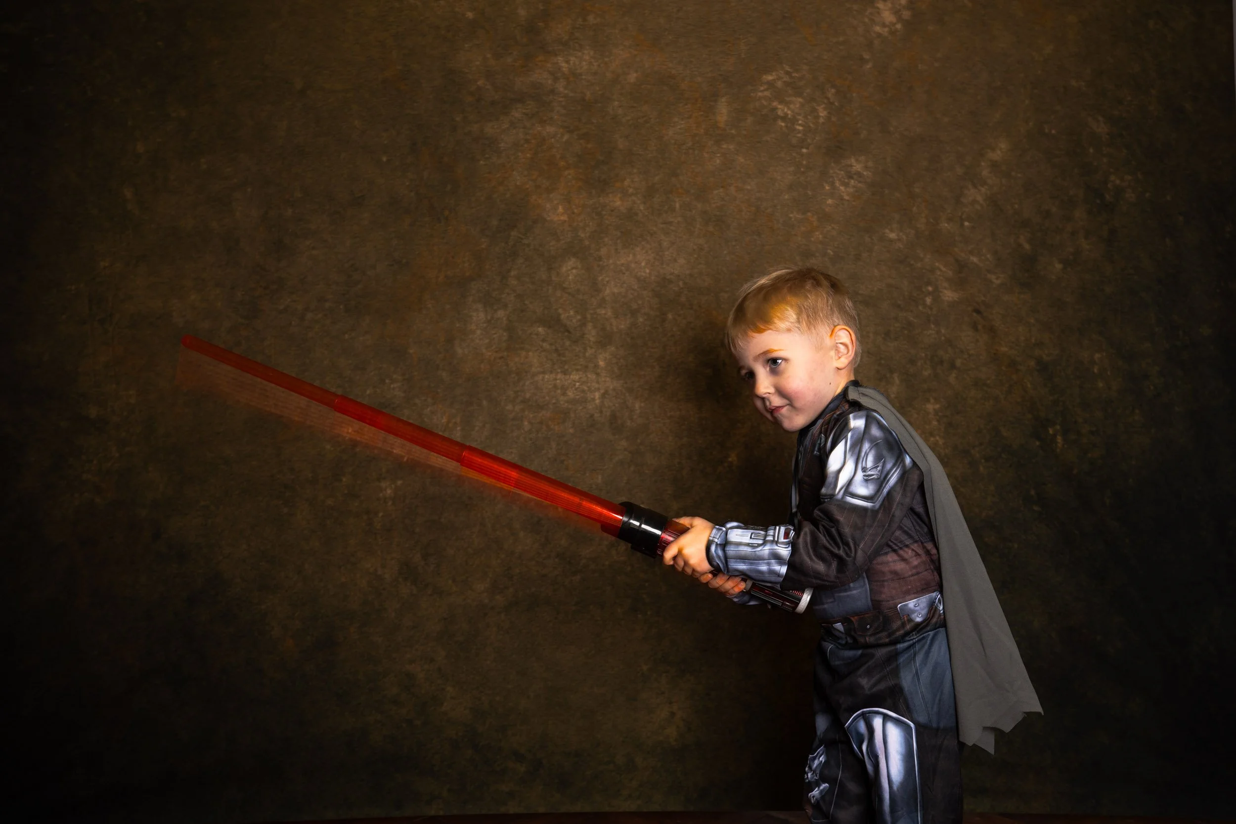 A young boy dressed in a Darth Vader costume, holding a red toy lightsaber, standing against a dark textured background.