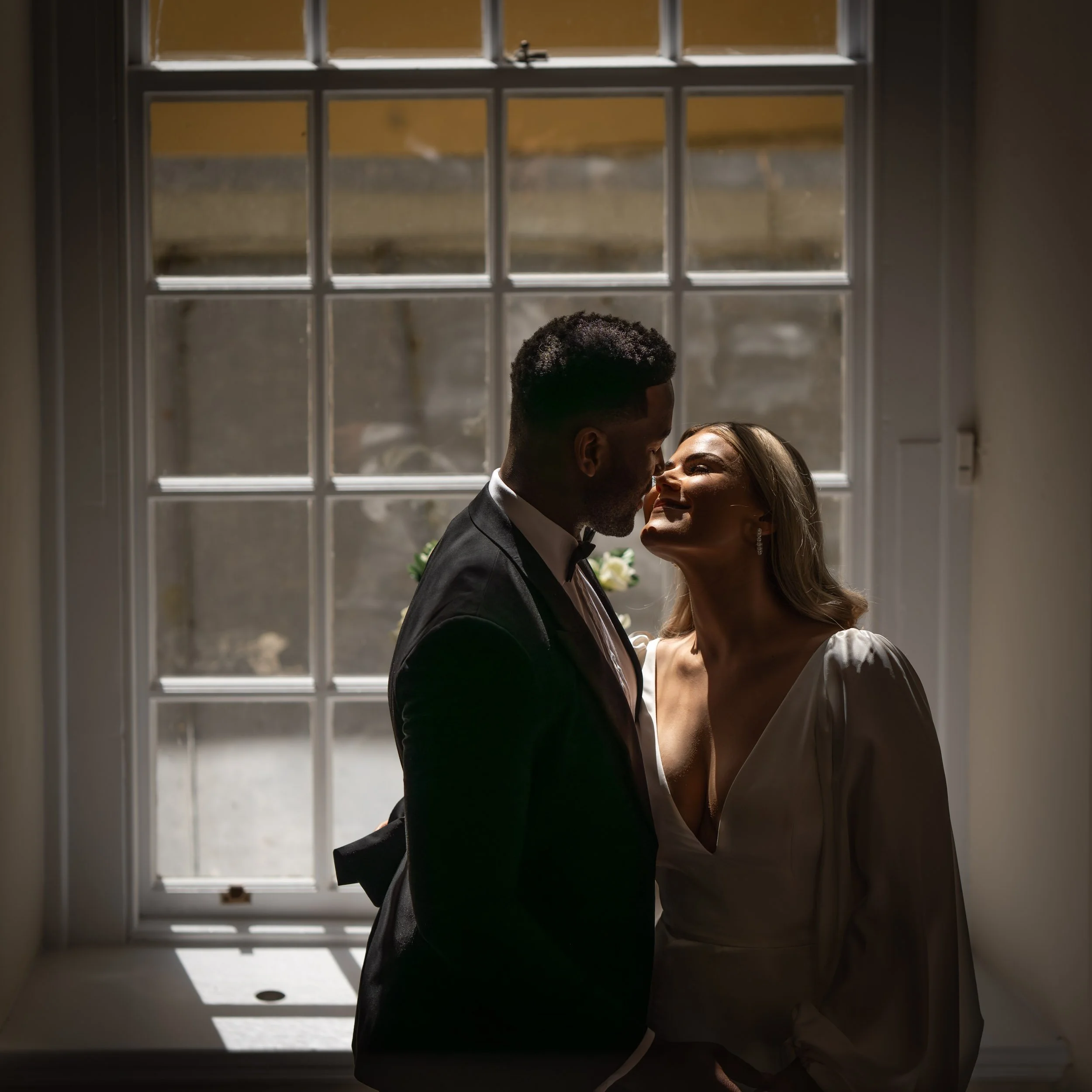 A couple about to kiss, standing close by a large window, with natural light creating silhouettes and soft shadows.