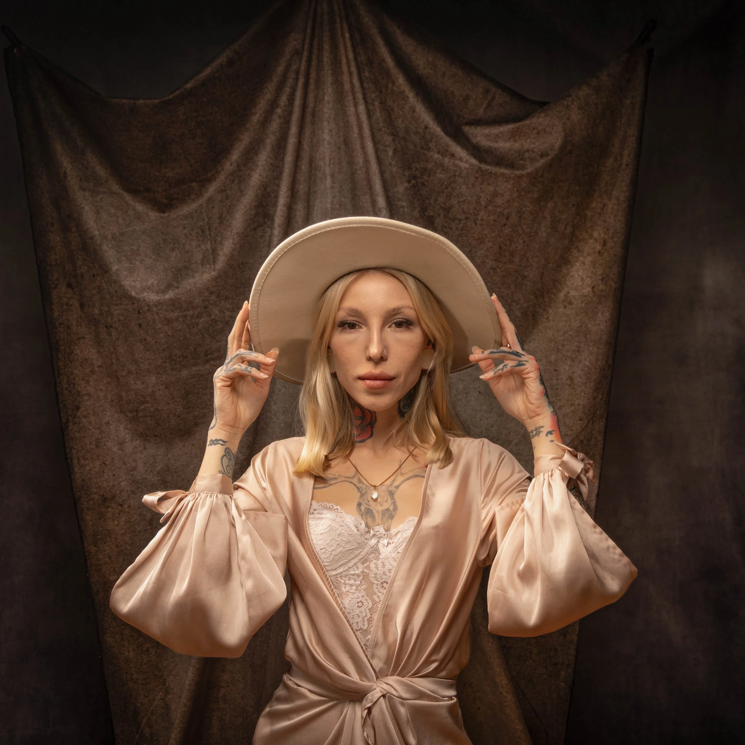 A woman with blonde hair, tattoos, and a neutral expression wears a beige wide-brimmed hat and a silky pink satin robe with lace details, standing in front of a textured brown backdrop.