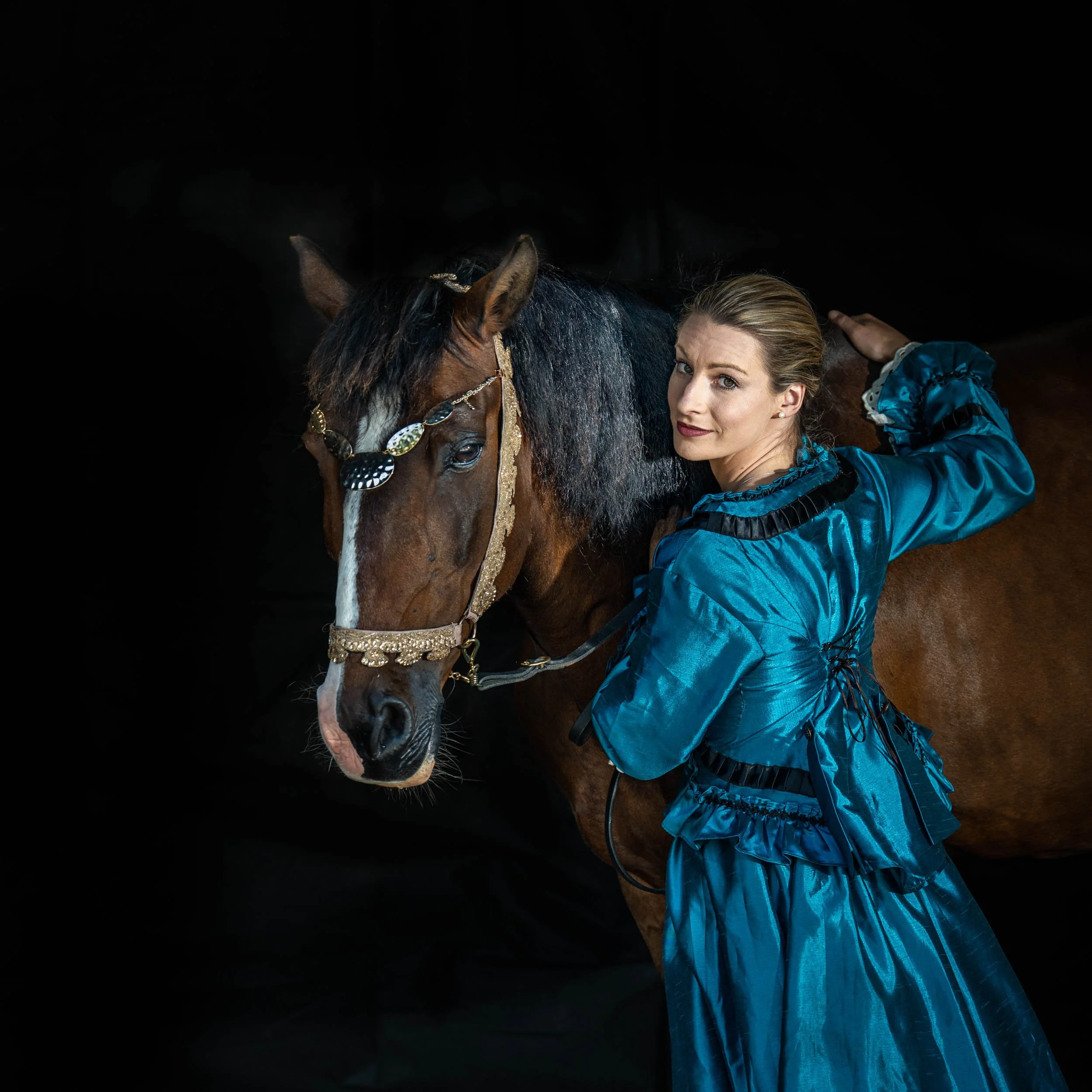 A woman in a blue dress standing next to a decorated brown horse against a black background.