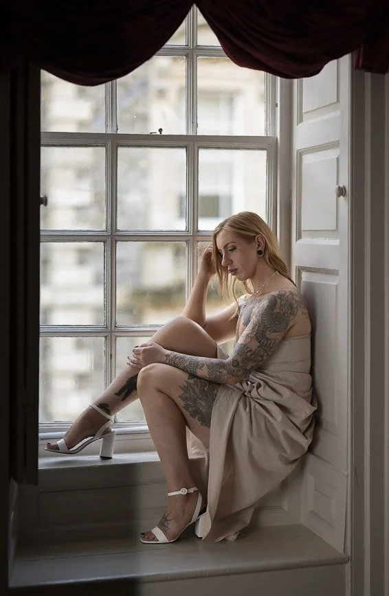 A woman with blonde hair, tattoos, and light skin, sitting on a window seat, wearing a beige dress and white high-heeled sandals, gazing down with her head resting on her hand, in a bright room with large window panels.