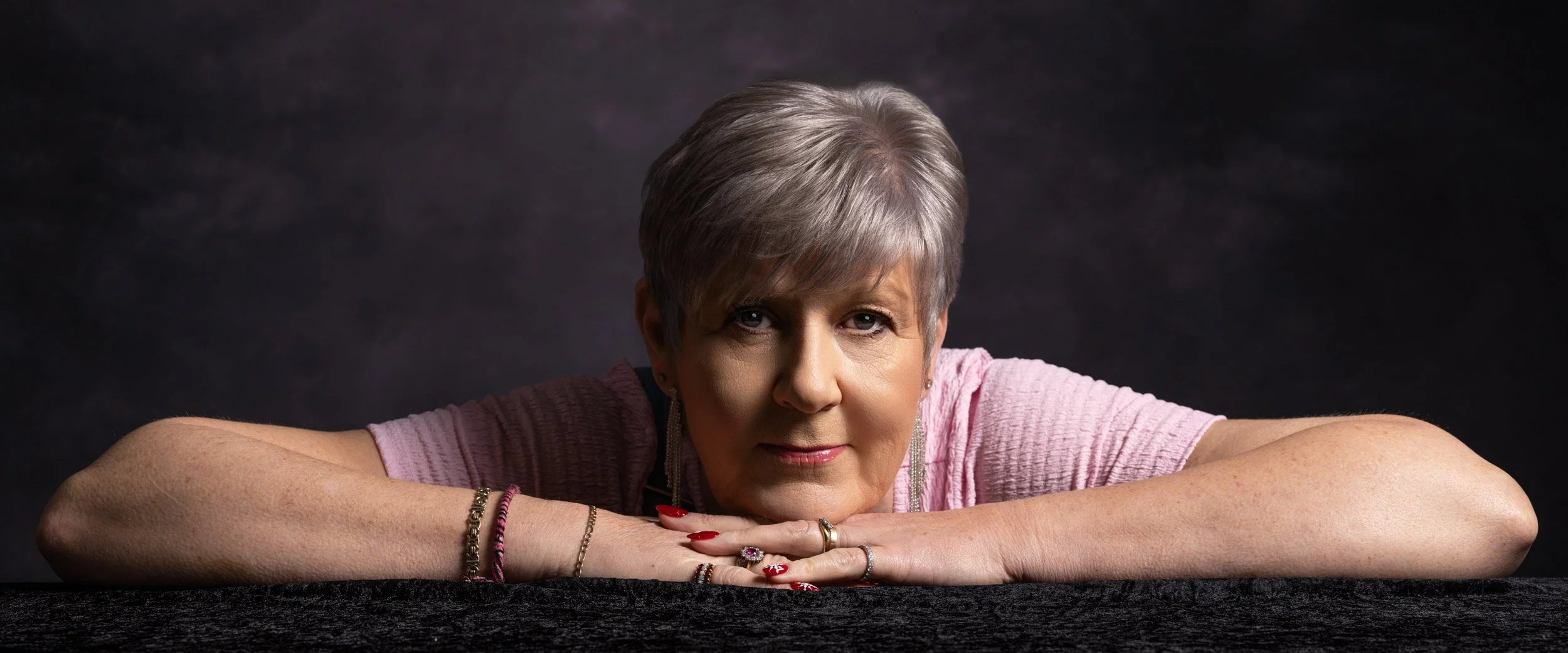 A woman with short gray hair leans on a dark surface with her arms crossed, looking directly at the camera with a neutral expression, wearing a pink top and jewelry, against a dark background.