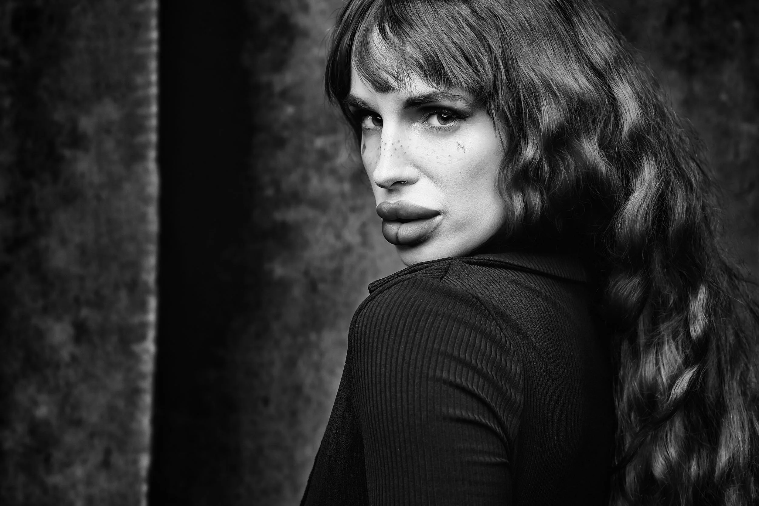 Black and white portrait of a woman with wavy hair, wearing a dark top, looking over her shoulder with a serious expression.