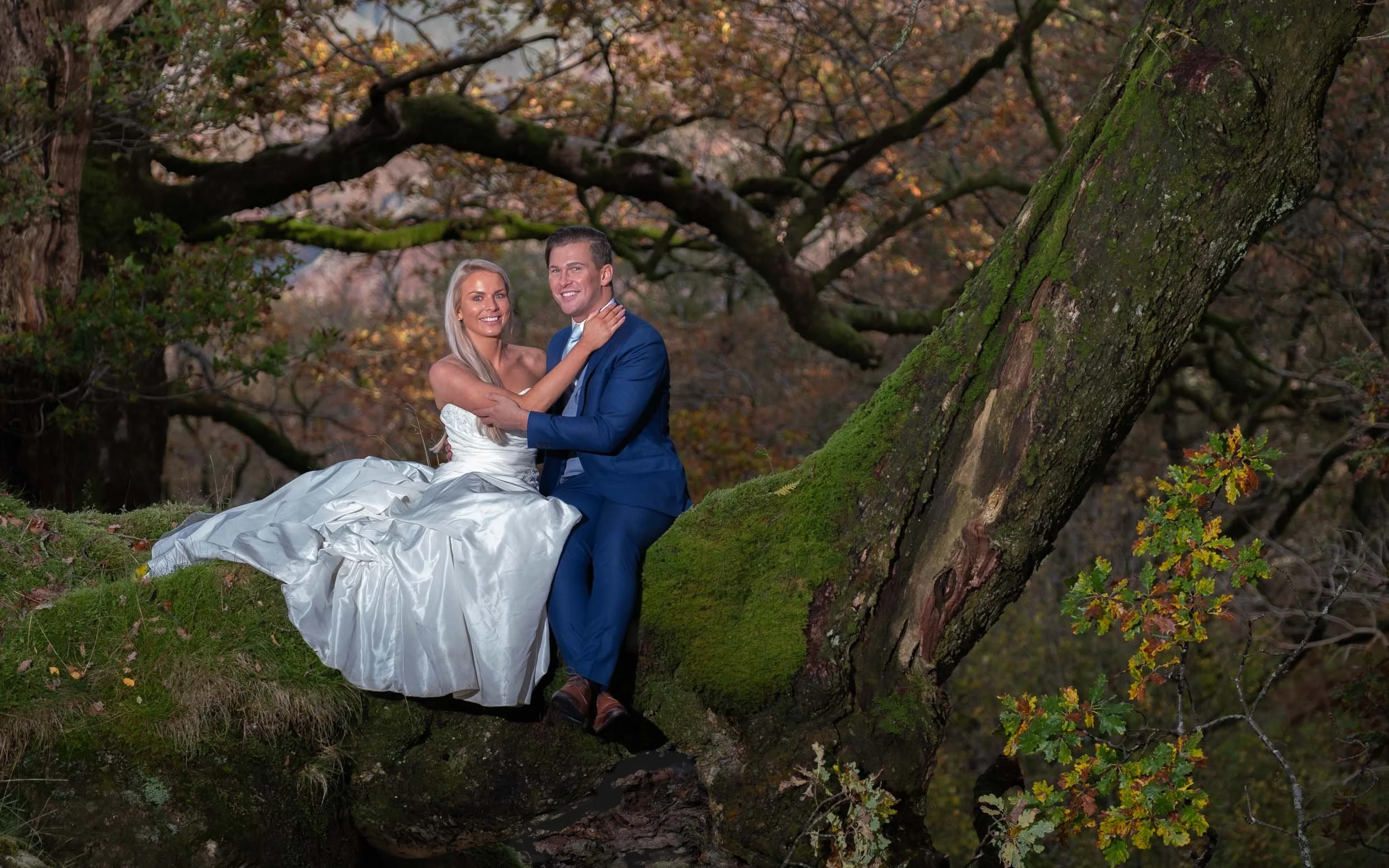 A smiling couple in wedding attire, the woman in a white gown and the man in a blue suit, sitting on a moss-covered tree branch in a forest with autumn leaves.