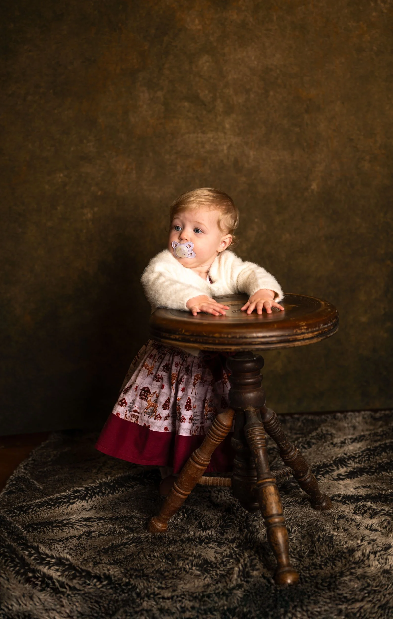 A young child with a pacifier in their mouth, wearing a cream-colored sweater and a patterned skirt, leaning on a wooden stool against a brown textured background.