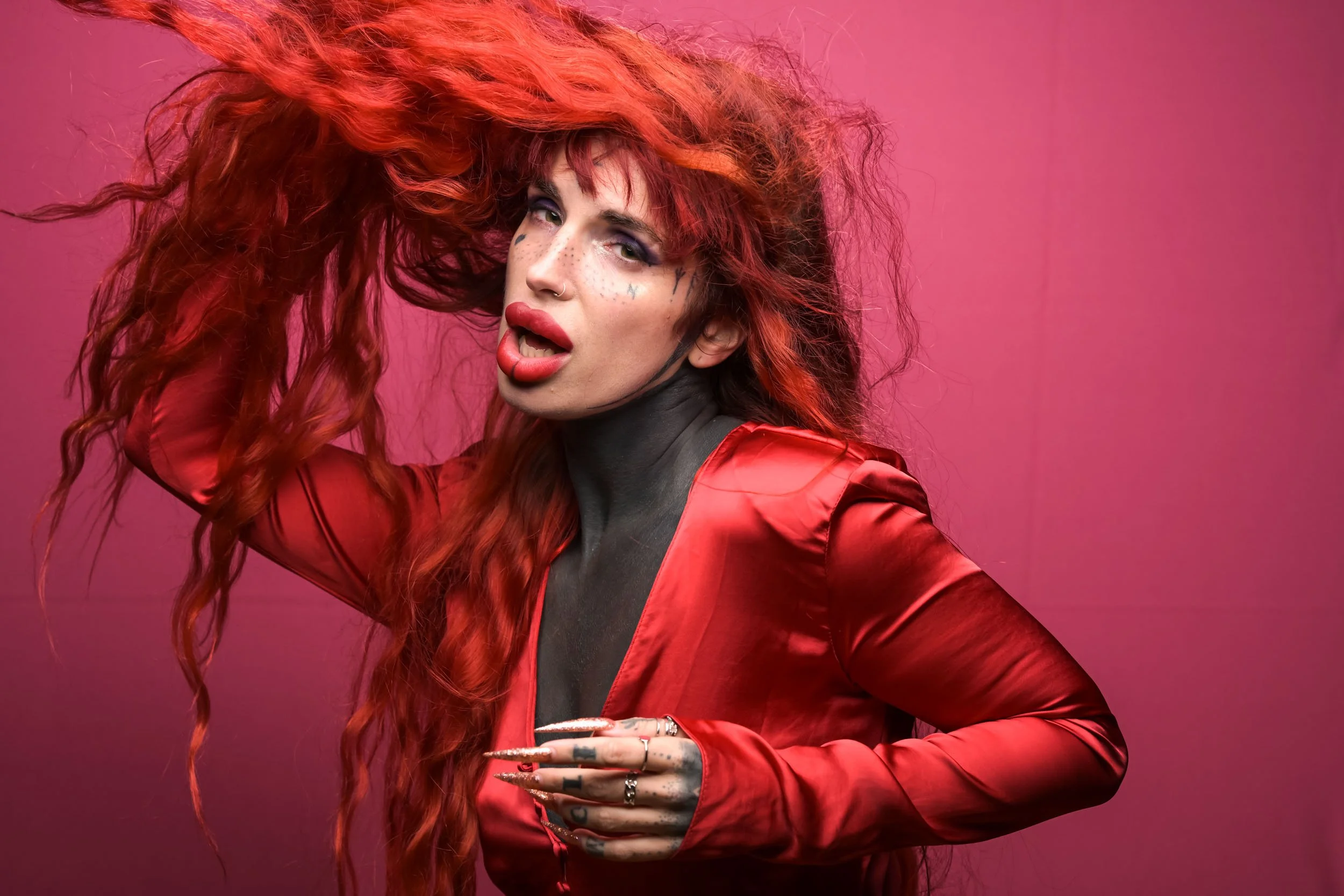 A woman with long, wavy red hair, wearing bright red lipstick, a red satin jacket, and tattoos on her hand, posing against a pink background.