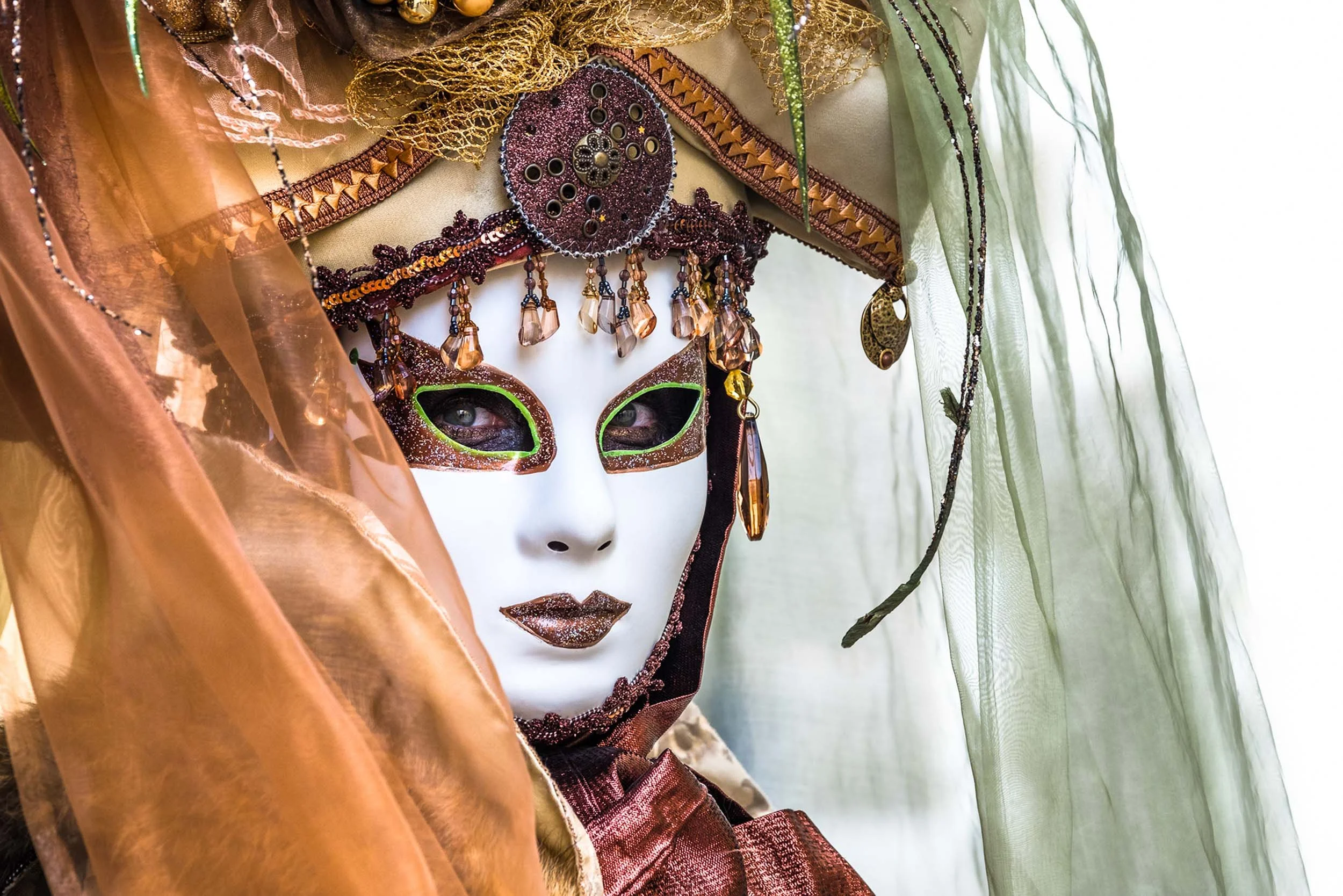 A person wearing a detailed, ornate mask with beads and jewels, surrounded by colorful fabrics and sheer green curtains.