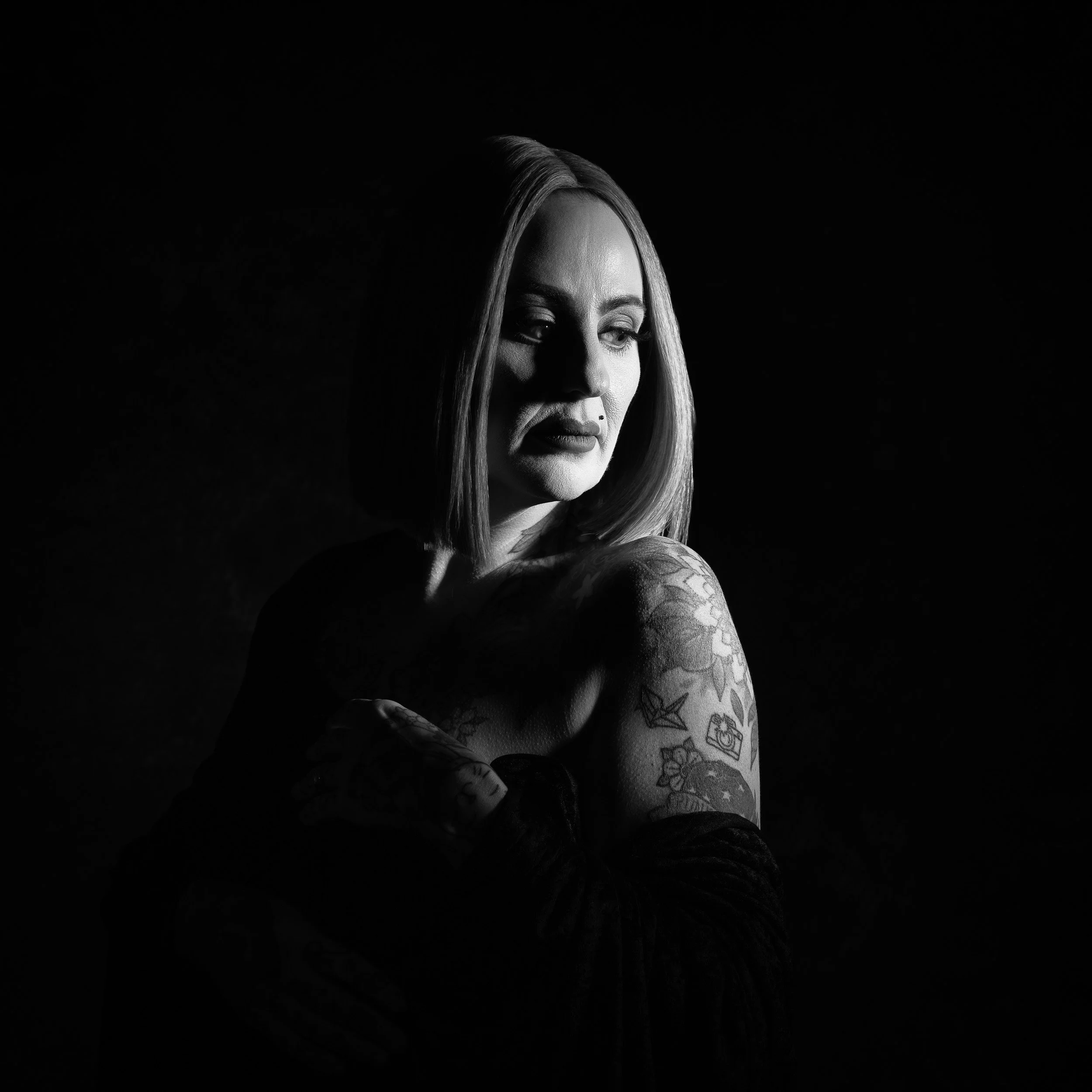Black and white portrait of a woman with tattoos, looking to the side with a serious expression, arms crossed, against a dark background.