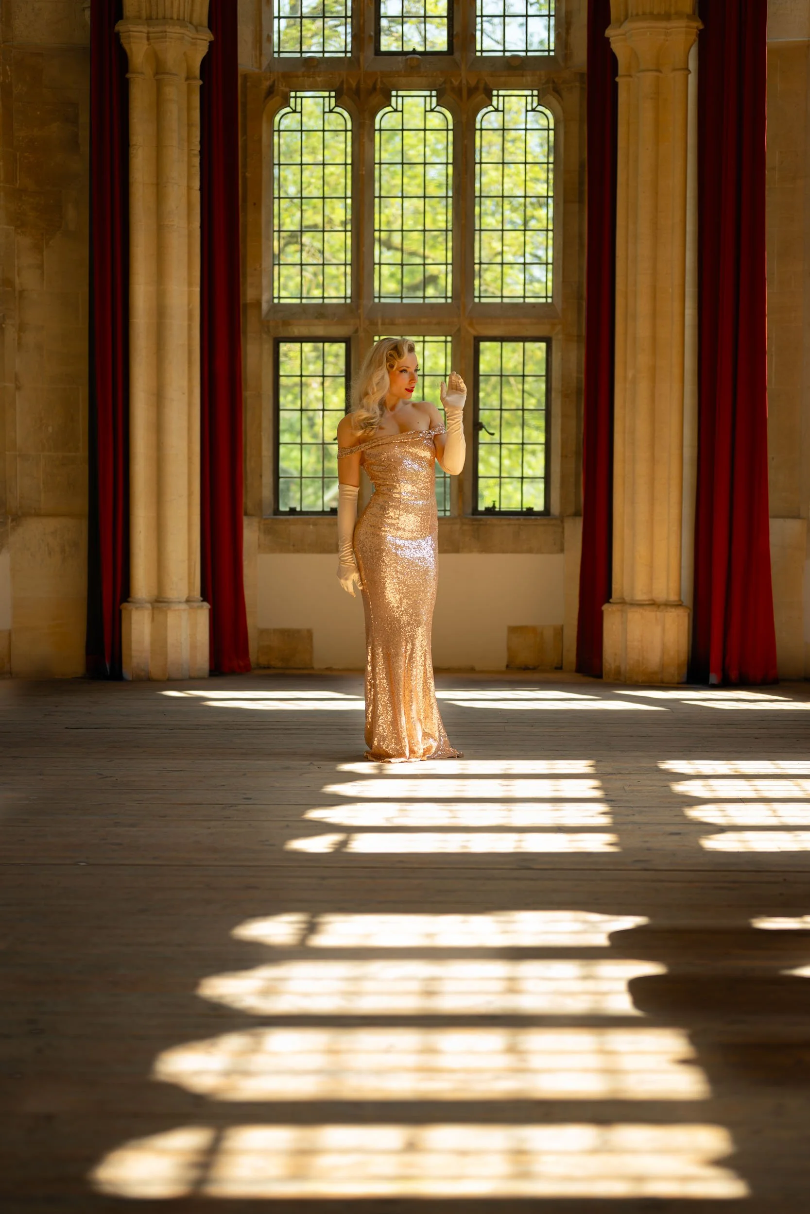 A woman in a gold sequined gown and long gloves standing in a sunlit hall with large stained glass windows and red curtains.