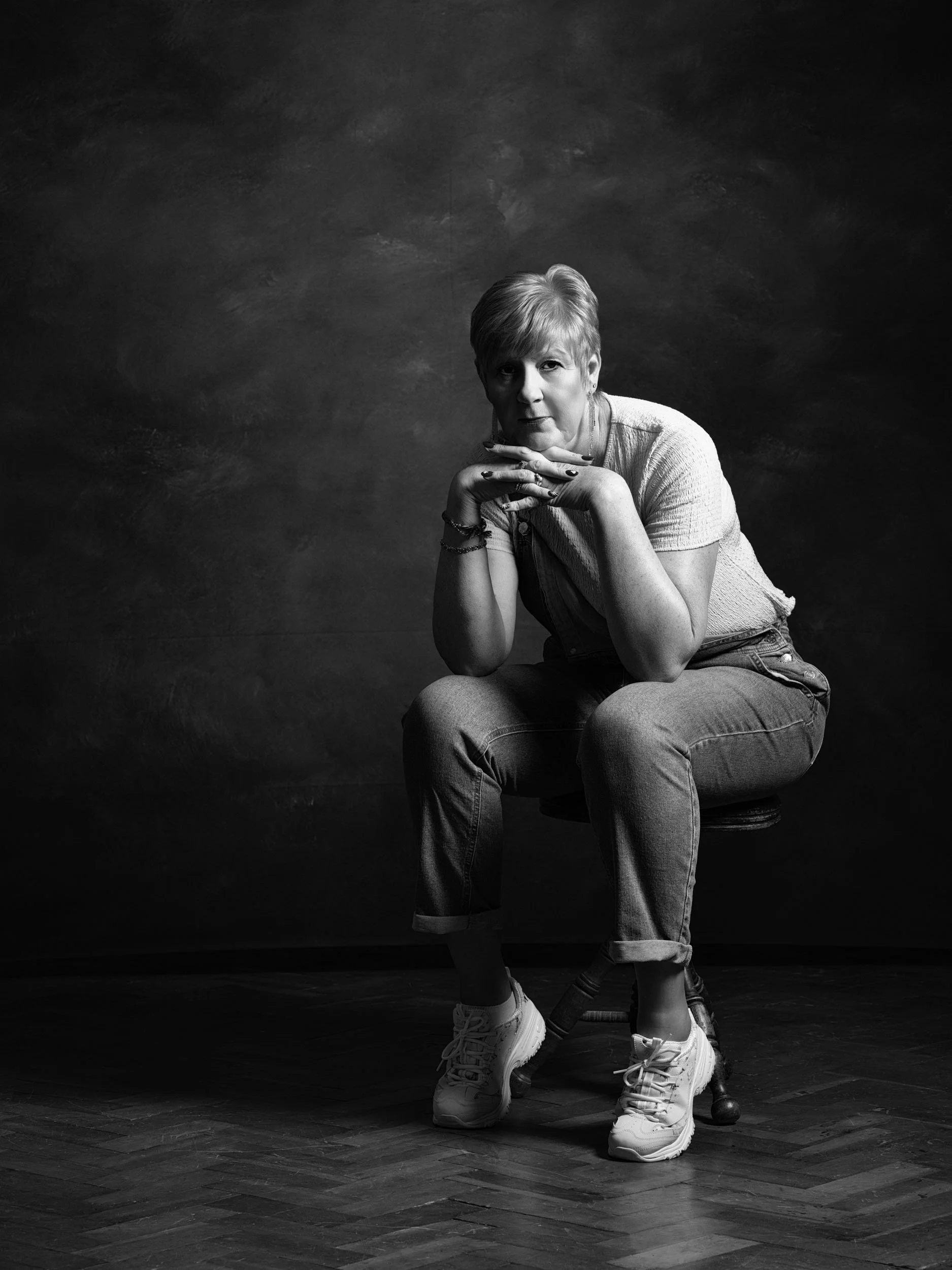 A woman with short hair sitting on a stool with her legs crossed and her hands resting under her chin, looking directly at the camera, in a black and white portrait.