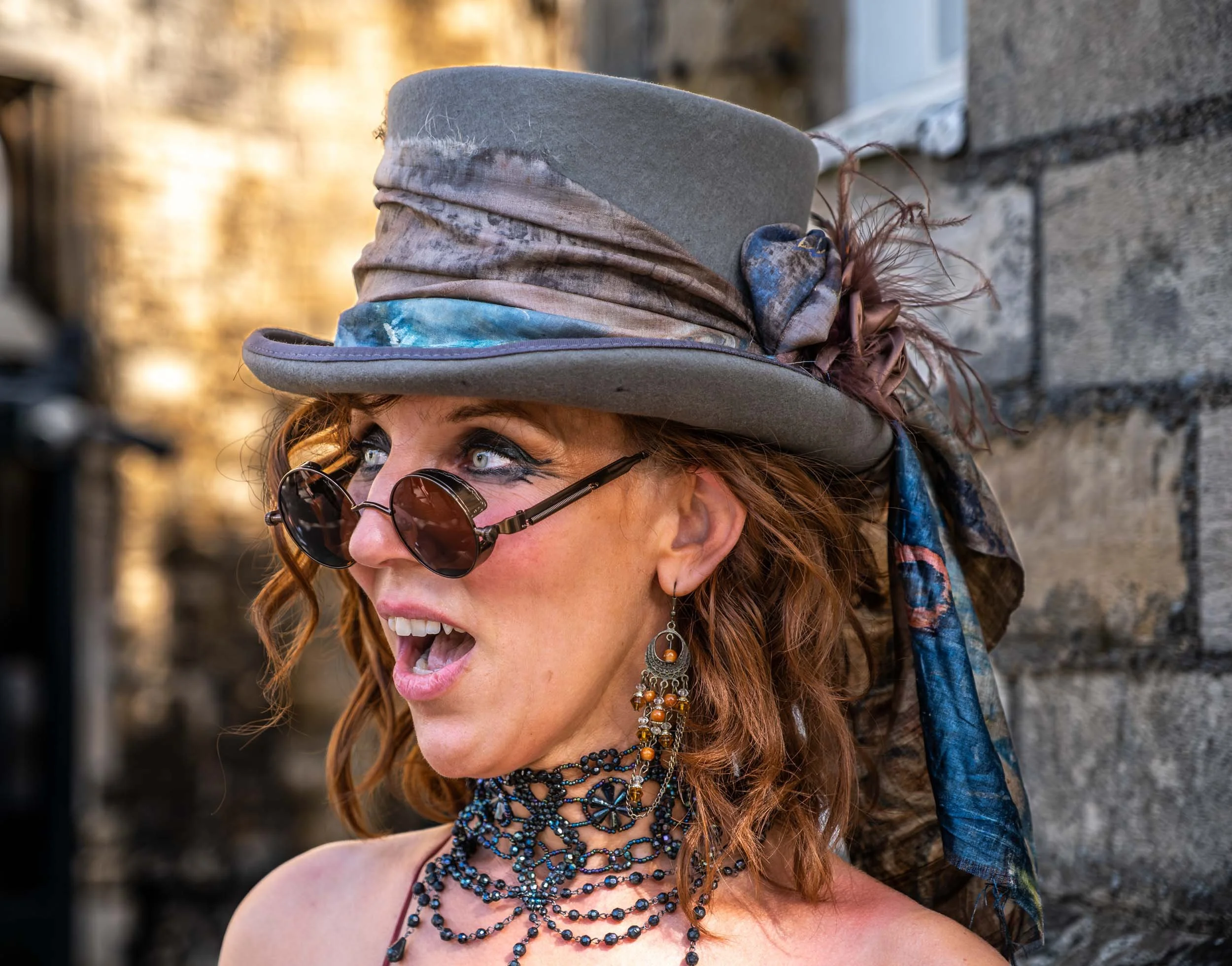 A woman with curly red hair wearing a gray hat with a colorful scarf, round sunglasses, and multiple layered necklaces, standing outdoors near a stone wall, looking to her right with her mouth open.