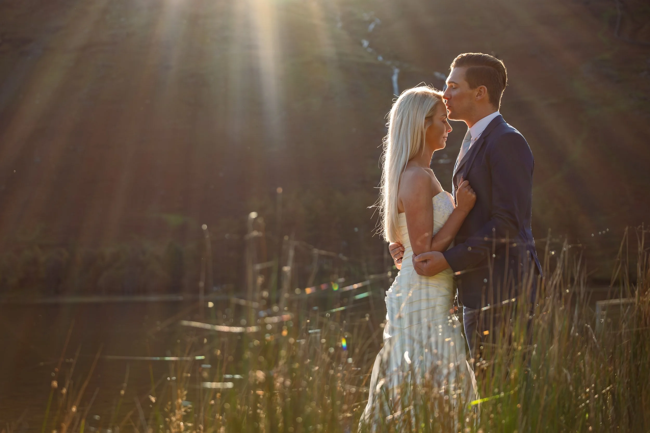 A couple in wedding attire standing close together outdoors during sunset, with a scenic background of a mountain and a waterfall.