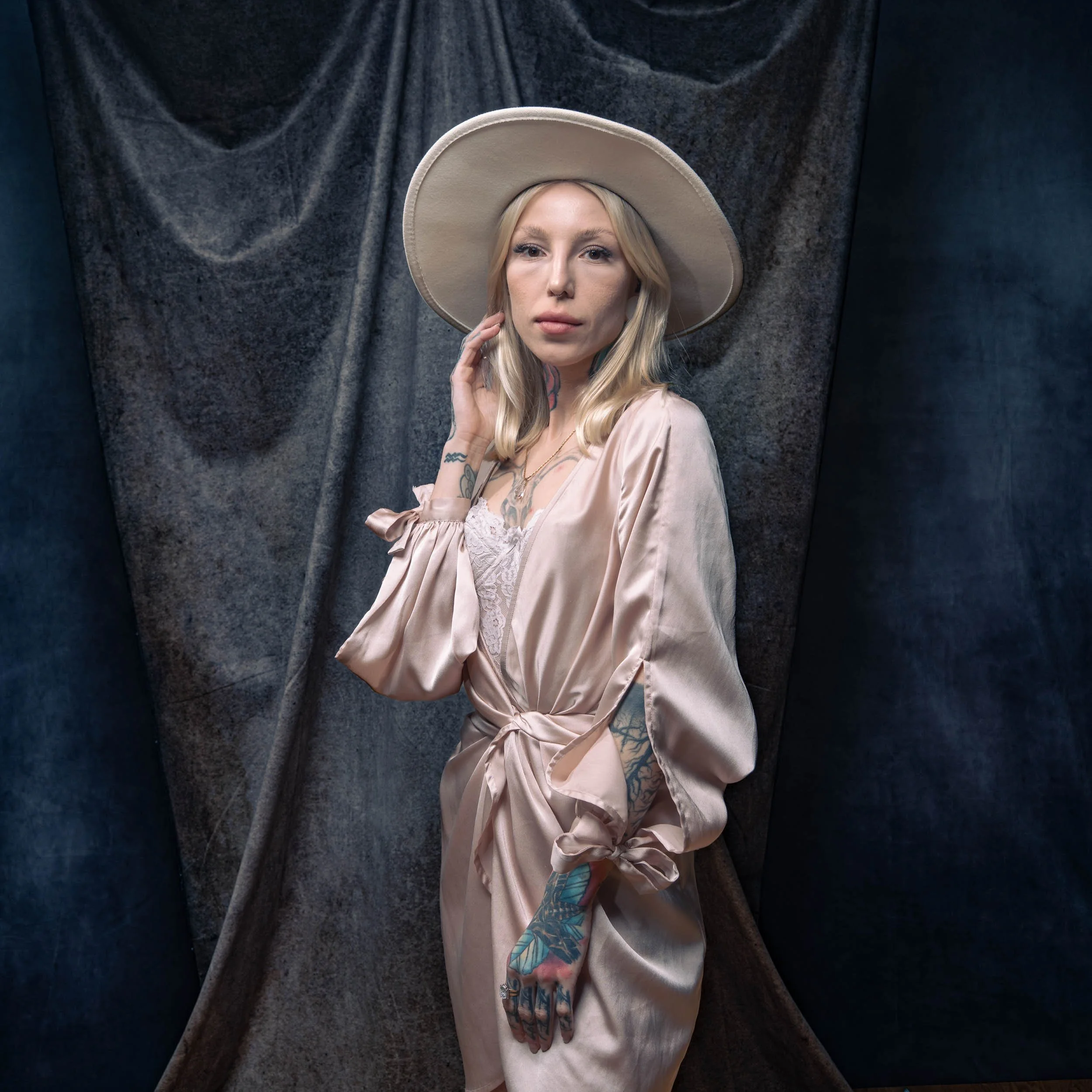 A woman with blonde hair wearing a wide-brimmed hat, satin robe, and lace top, standing against a dark textured background.