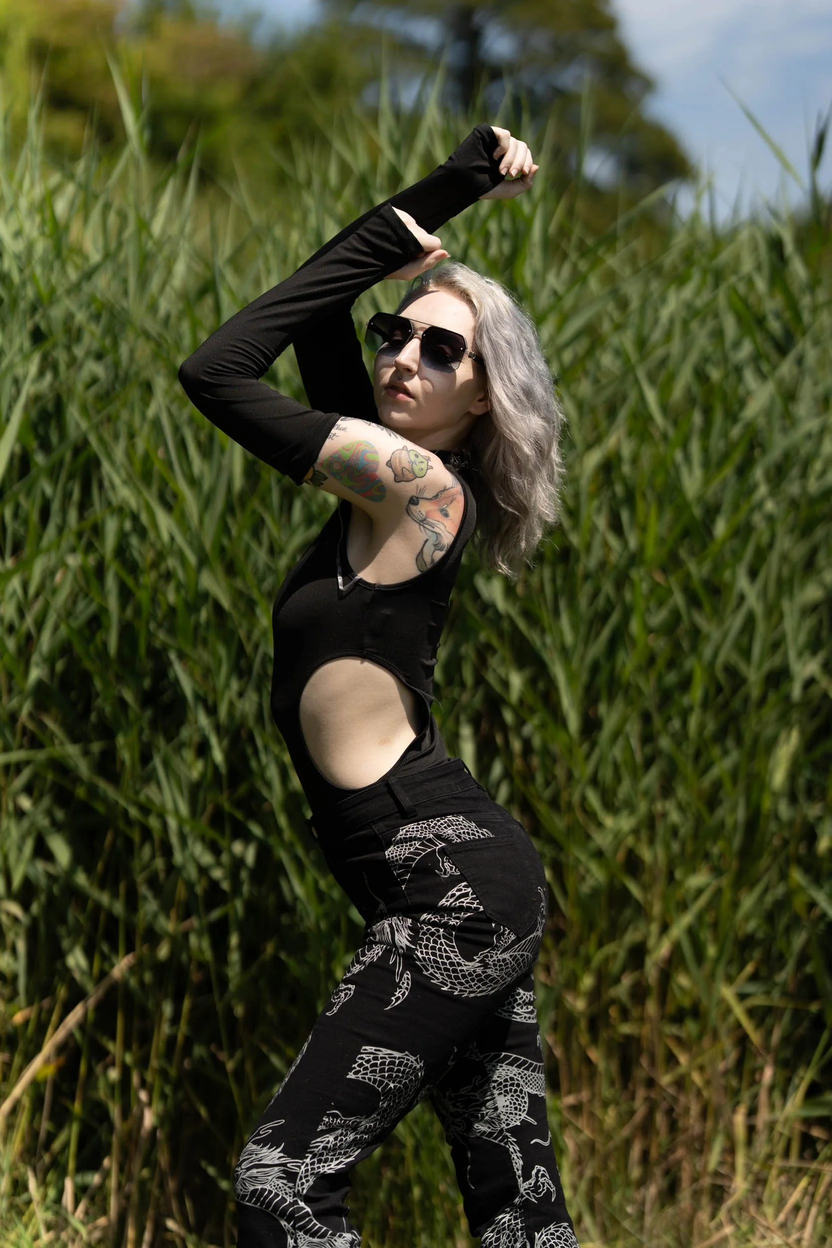 A woman with wavy silver hair and tattoos on her arm stands in front of tall green plants, wearing black sunglasses, a black sleeveless top with cutouts, and black pants with white dragon designs.