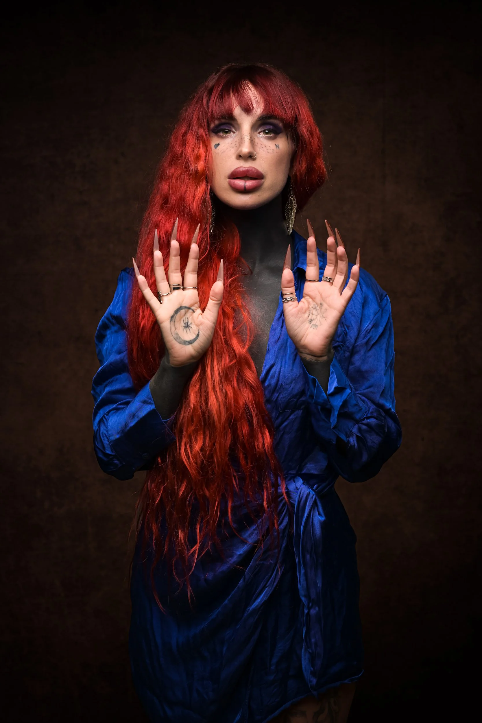 A woman with long, wavy red hair and tattoos on her hands and neck, wearing a blue dress, with makeup including bold eyeshadow, lipstick, and small face tattoos, poses against a dark background.