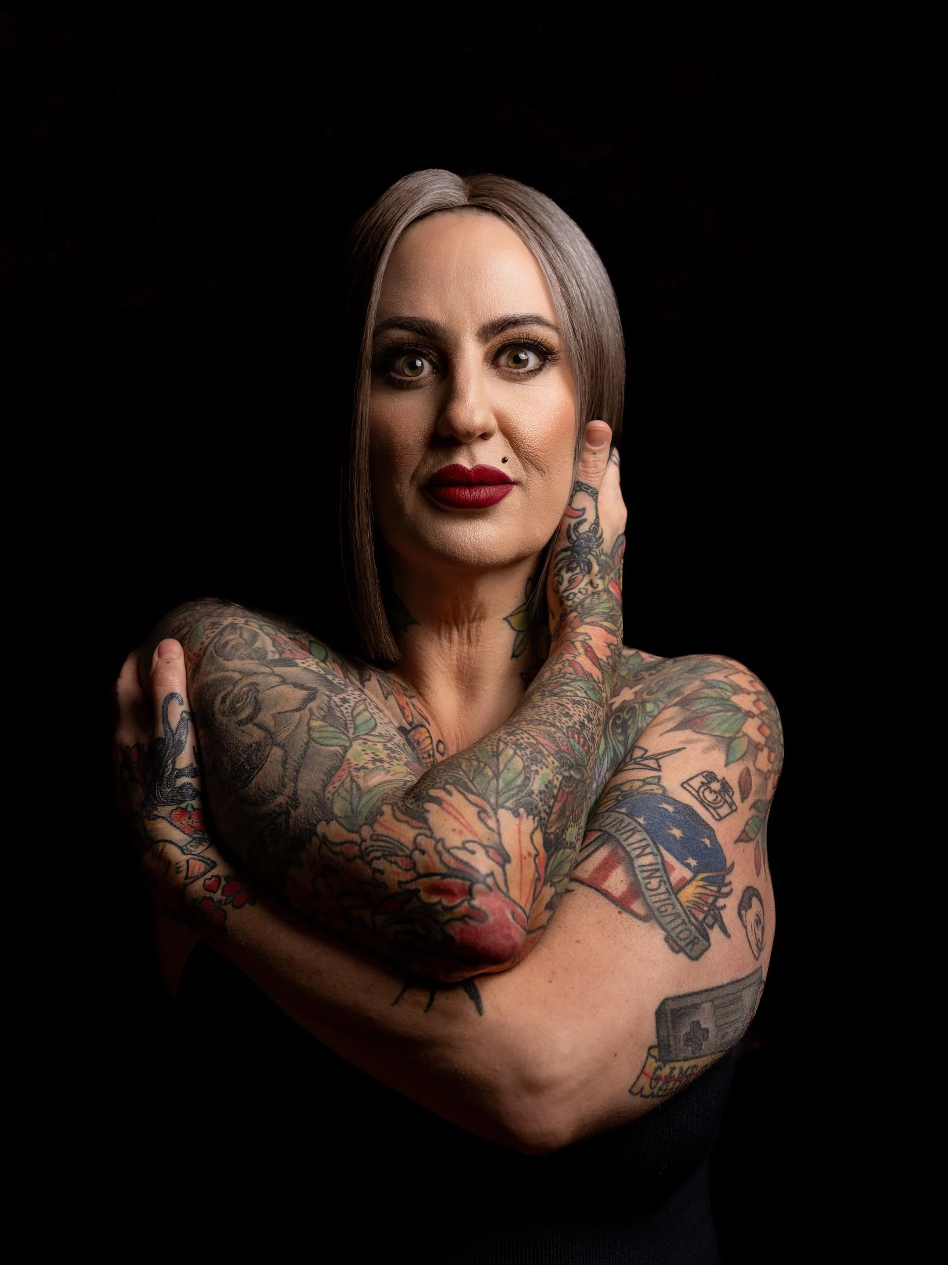 A woman with shoulder-length dark hair, bold makeup, and extensive colorful tattoos on her arms and shoulders, posing with her hands near her face against a black background.