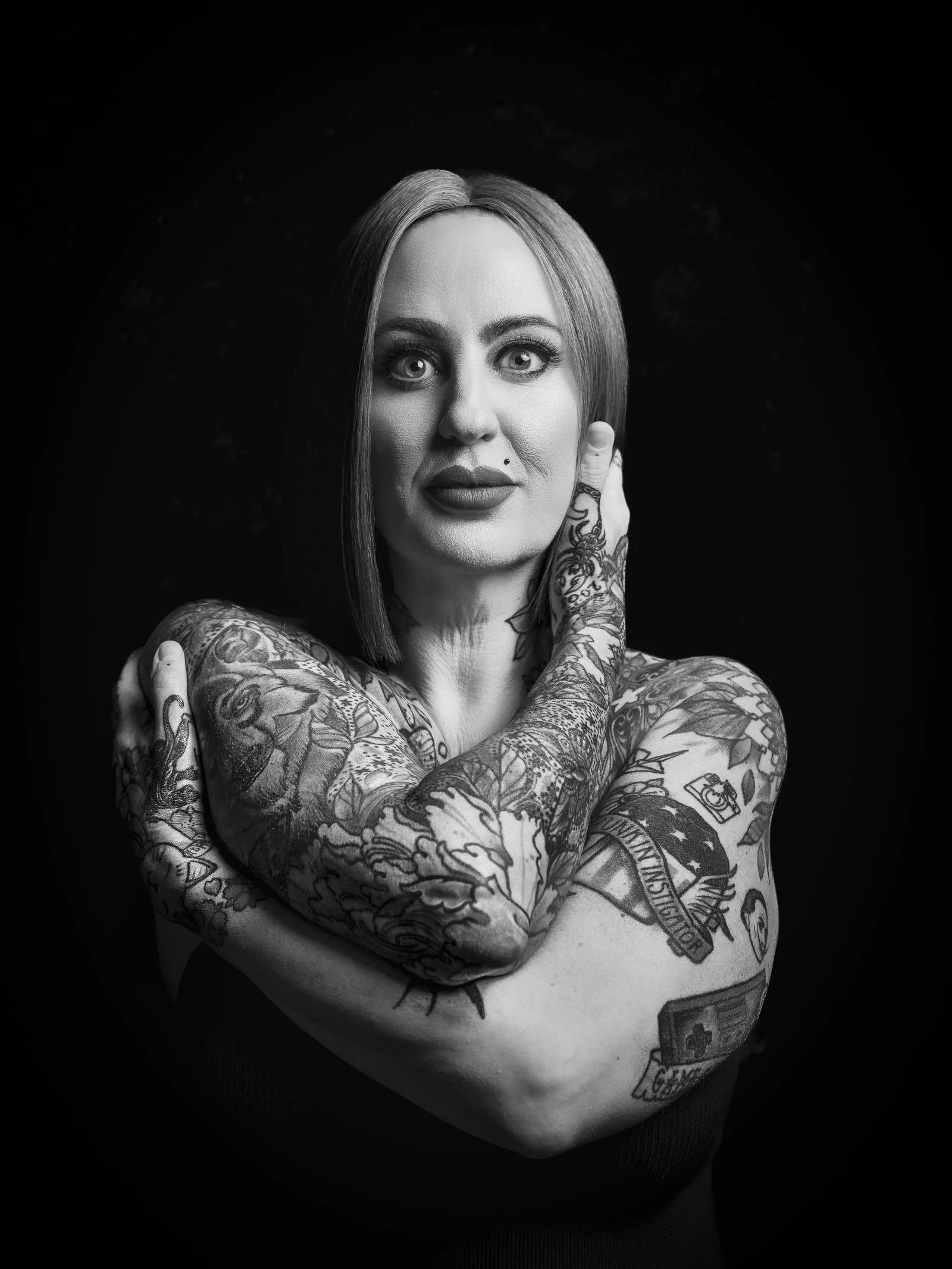 Black and white portrait of a woman with shoulder-length hair, heavily tattooed arms, and expressive makeup, looking directly at the camera with her left hand on the side of her face.