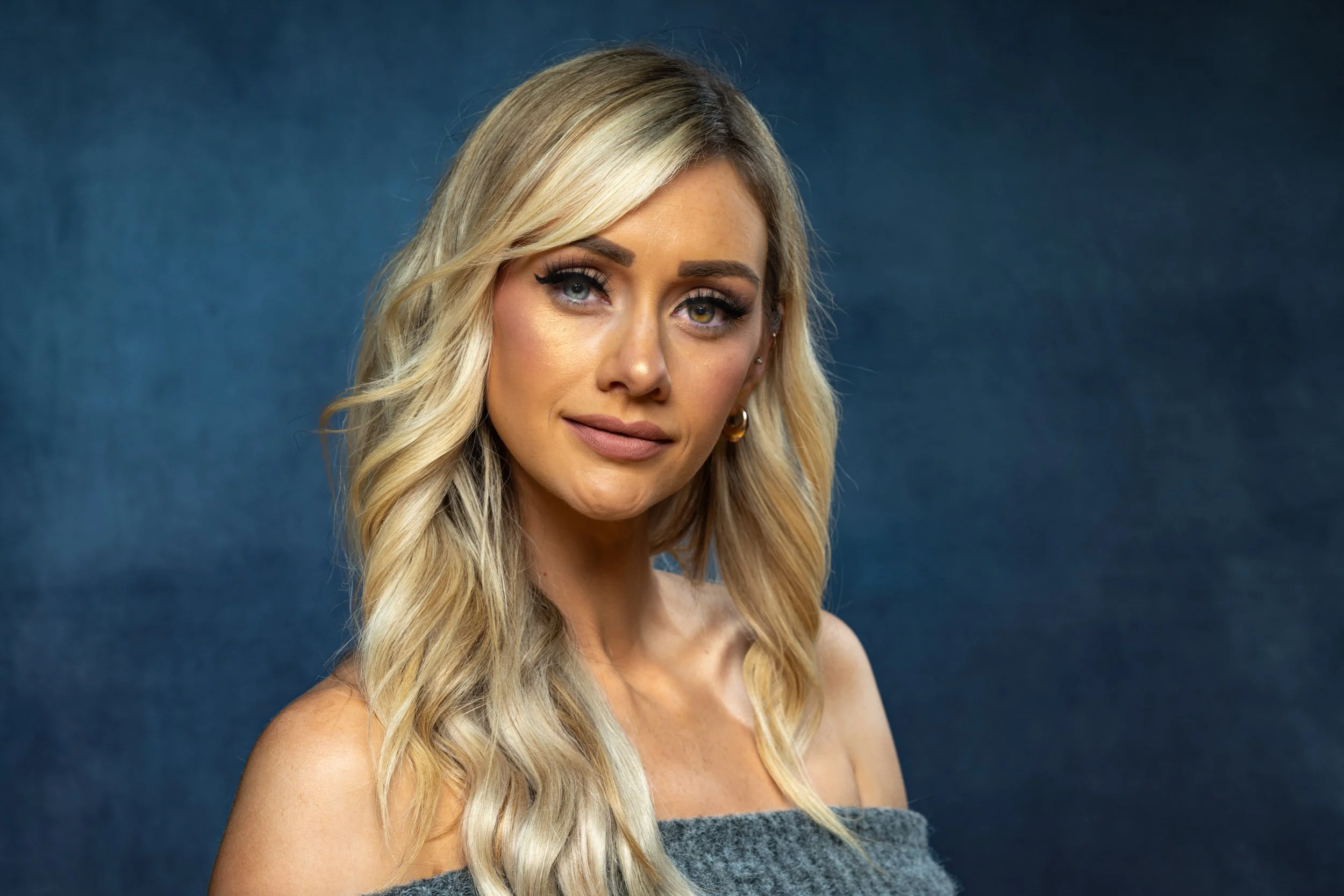 A woman with long blonde hair, wearing makeup and gold hoop earrings, is posing against a dark blue background.