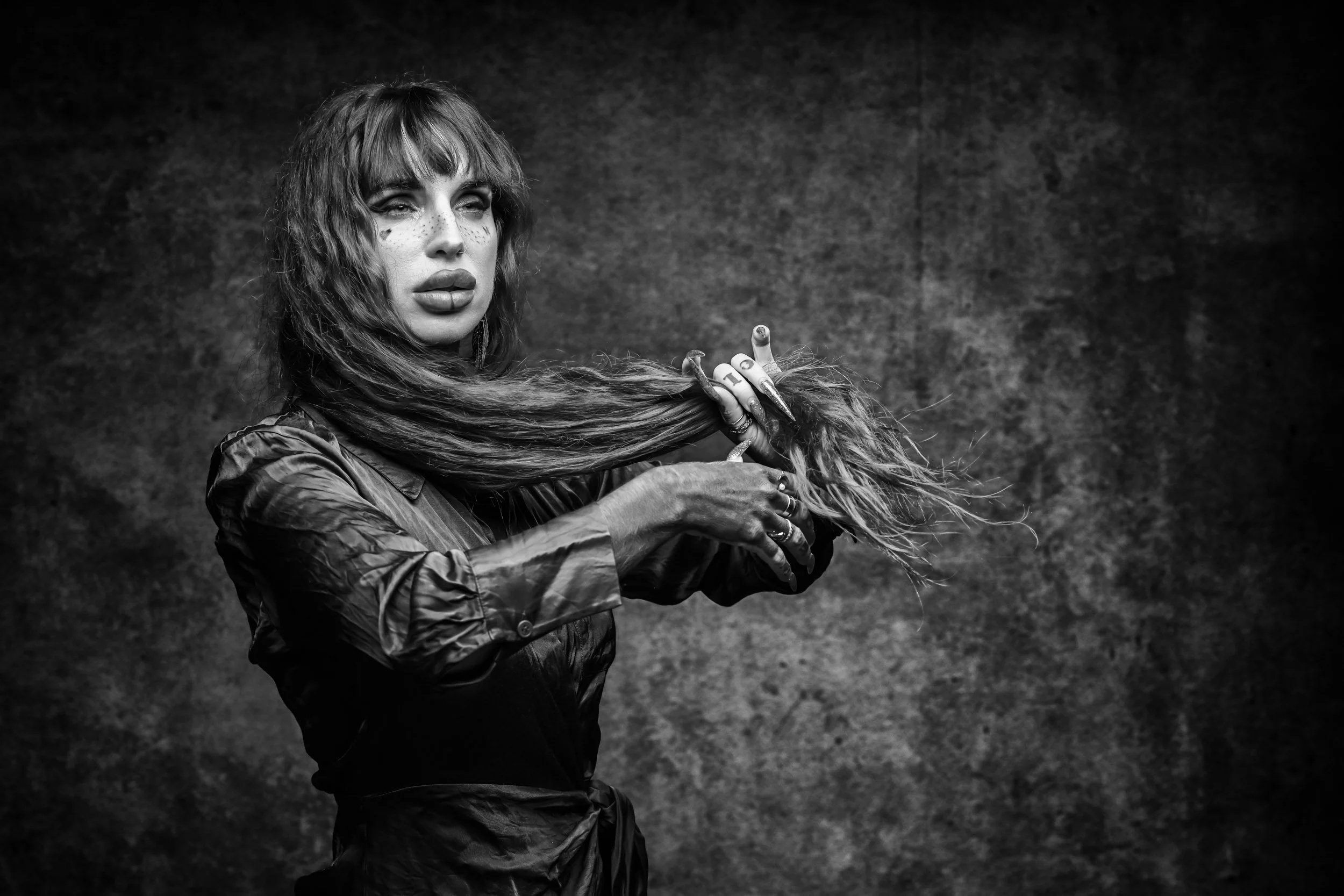 Black and white portrait of a woman with long, wavy hair, wearing a shiny, long-sleeved shirt, holding her hair in one hand, standing against a textured background.