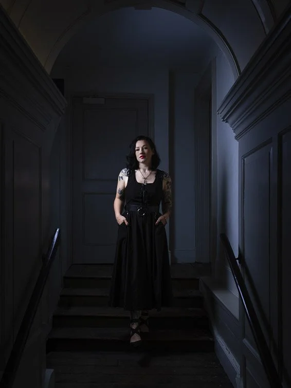 A woman with dark hair and tattoos wearing a black dress stands on a wooden staircase in a dimly lit hallway with dark blue walls and a rounded ceiling.