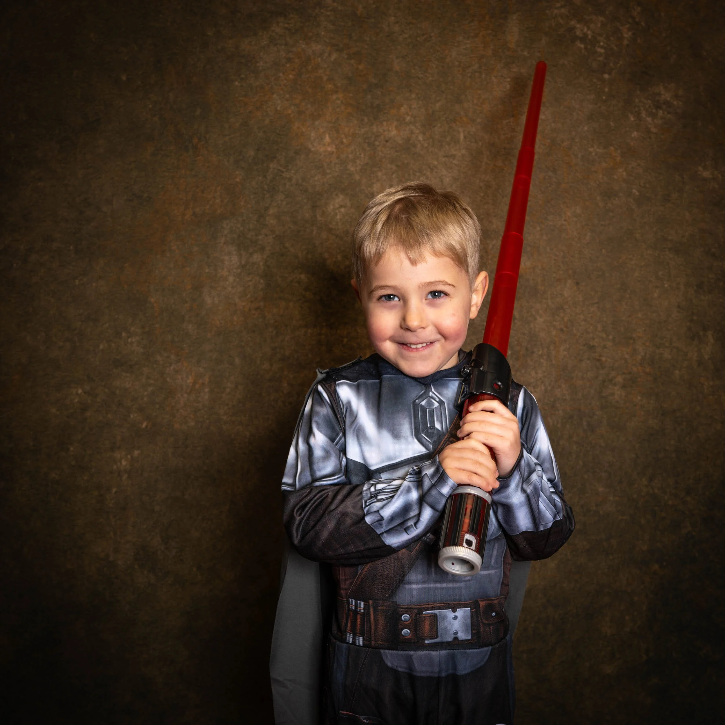 A young boy dressed as a Jedi from Star Wars, holding a red lightsaber, standing against a brown textured background, smiling at the camera.