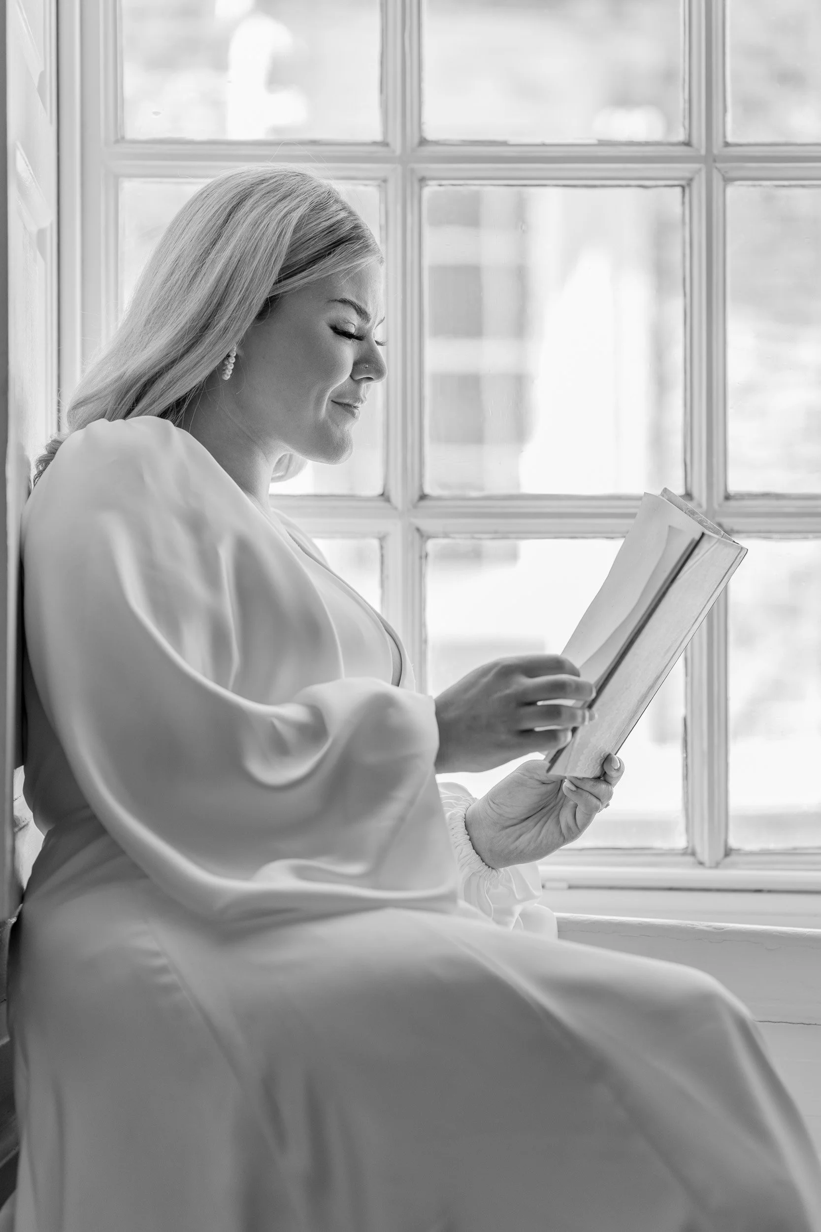 A woman with long blonde hair wearing earrings, sitting by a window and reading a book.