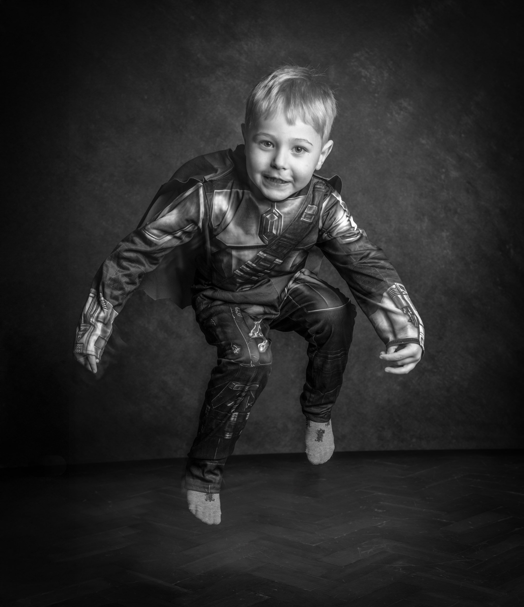 A young boy in a superhero costume appears to be jumping or crouching in a studio with a dark background.