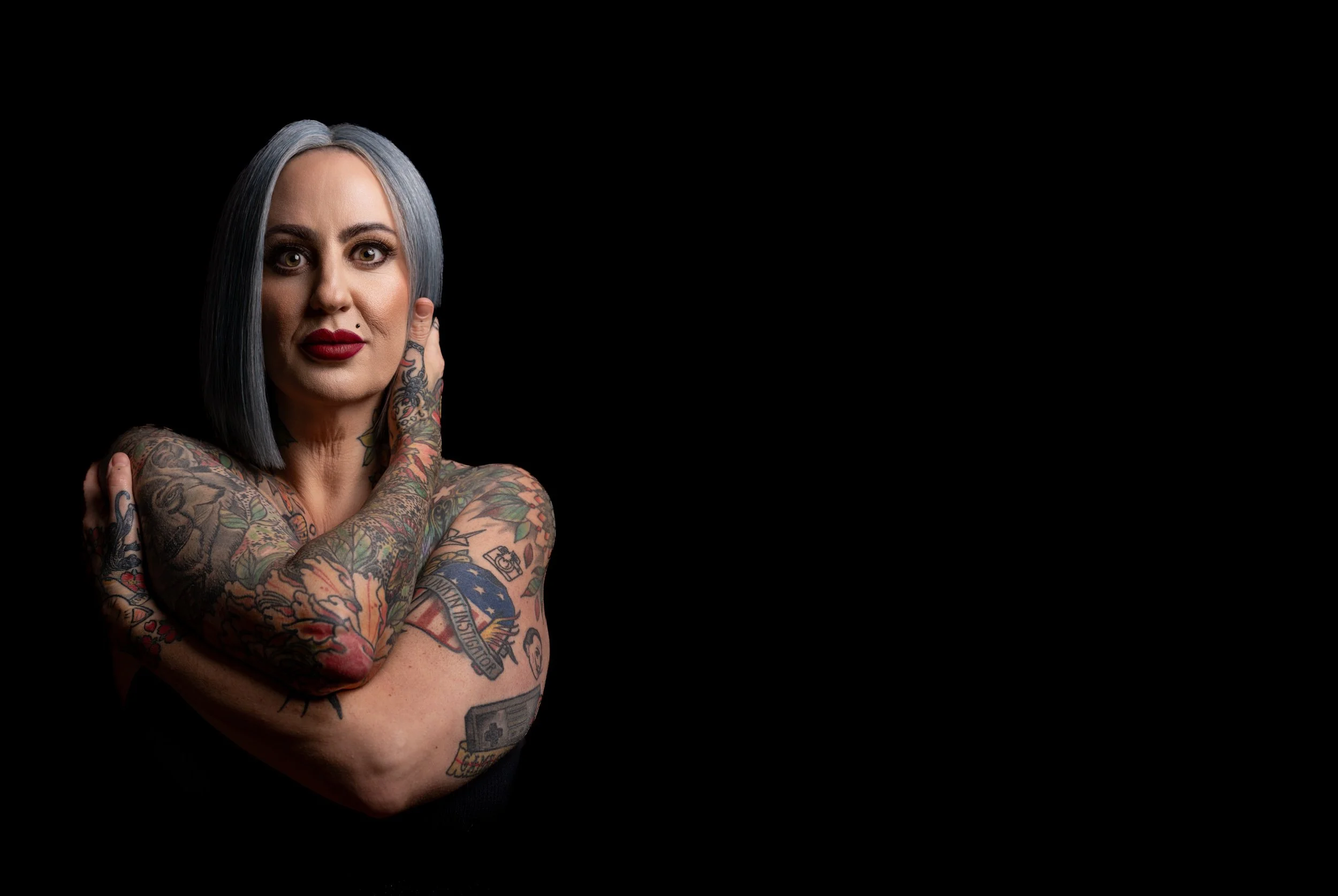 Tattooed woman with gray hair, red lipstick, and a confident expression poses against a black background.
