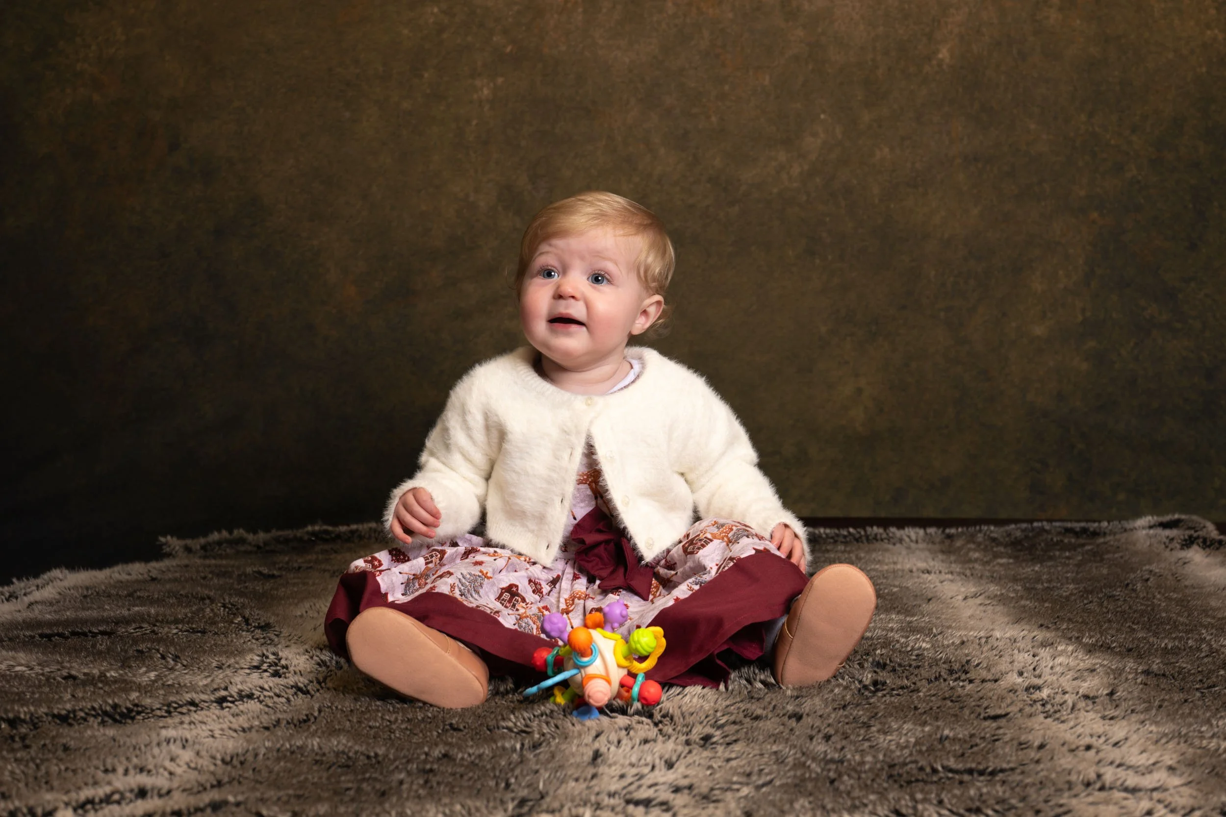 A baby girl sitting on a plush carpet, wearing a cream-colored cardigan and a patterned skirt, with colorful toys in front of her, looking up with a distressed expression.