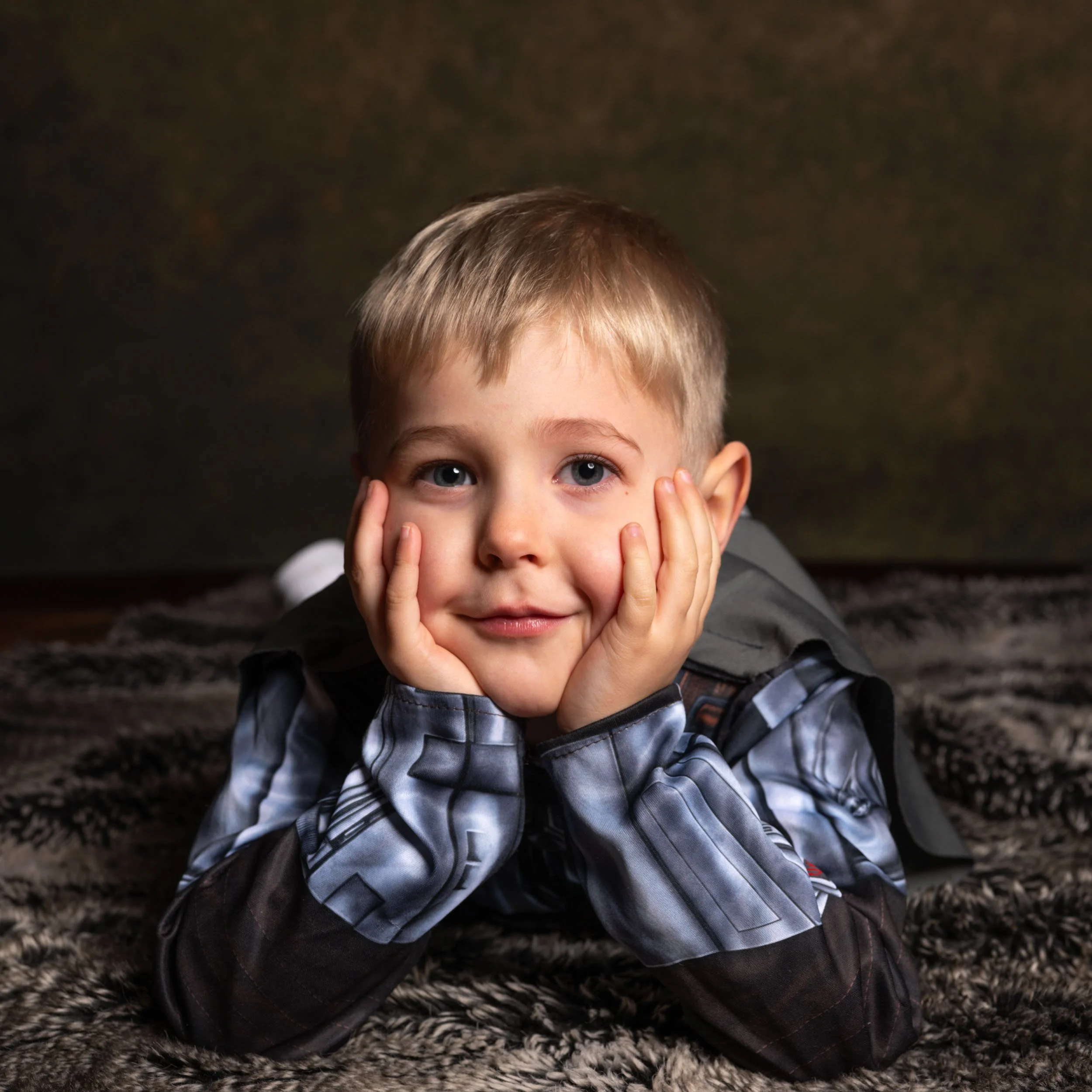 A young boy with blonde hair and blue eyes rests his face in his hands, lying on his stomach on a furry blanket, looking at the camera with a gentle smile.