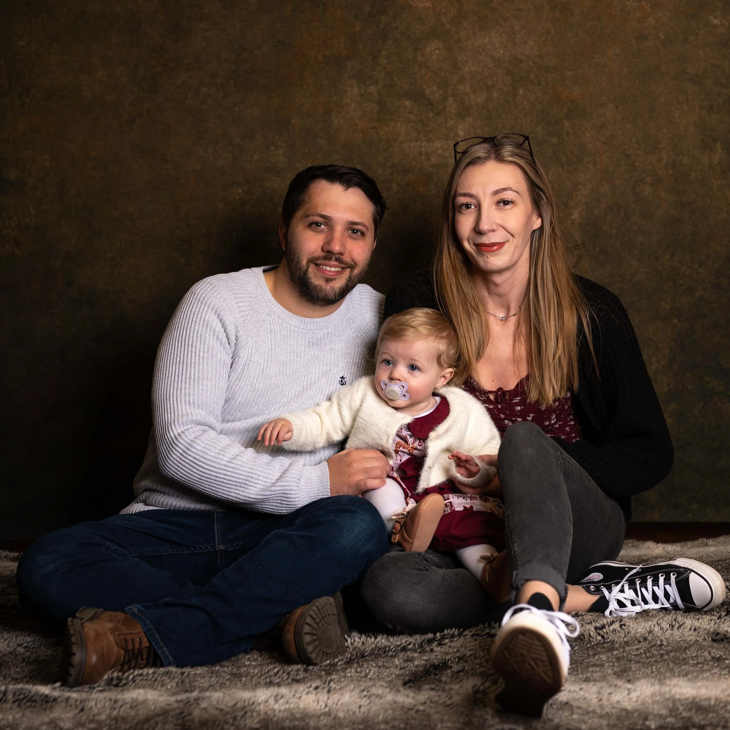 A family of three sitting on a rug against a brown background. The man has dark hair and a beard, wearing a light gray sweater. The woman has long light brown hair, glasses on her head, and is wearing a dark sweater and sneakers. The young girl, sitt