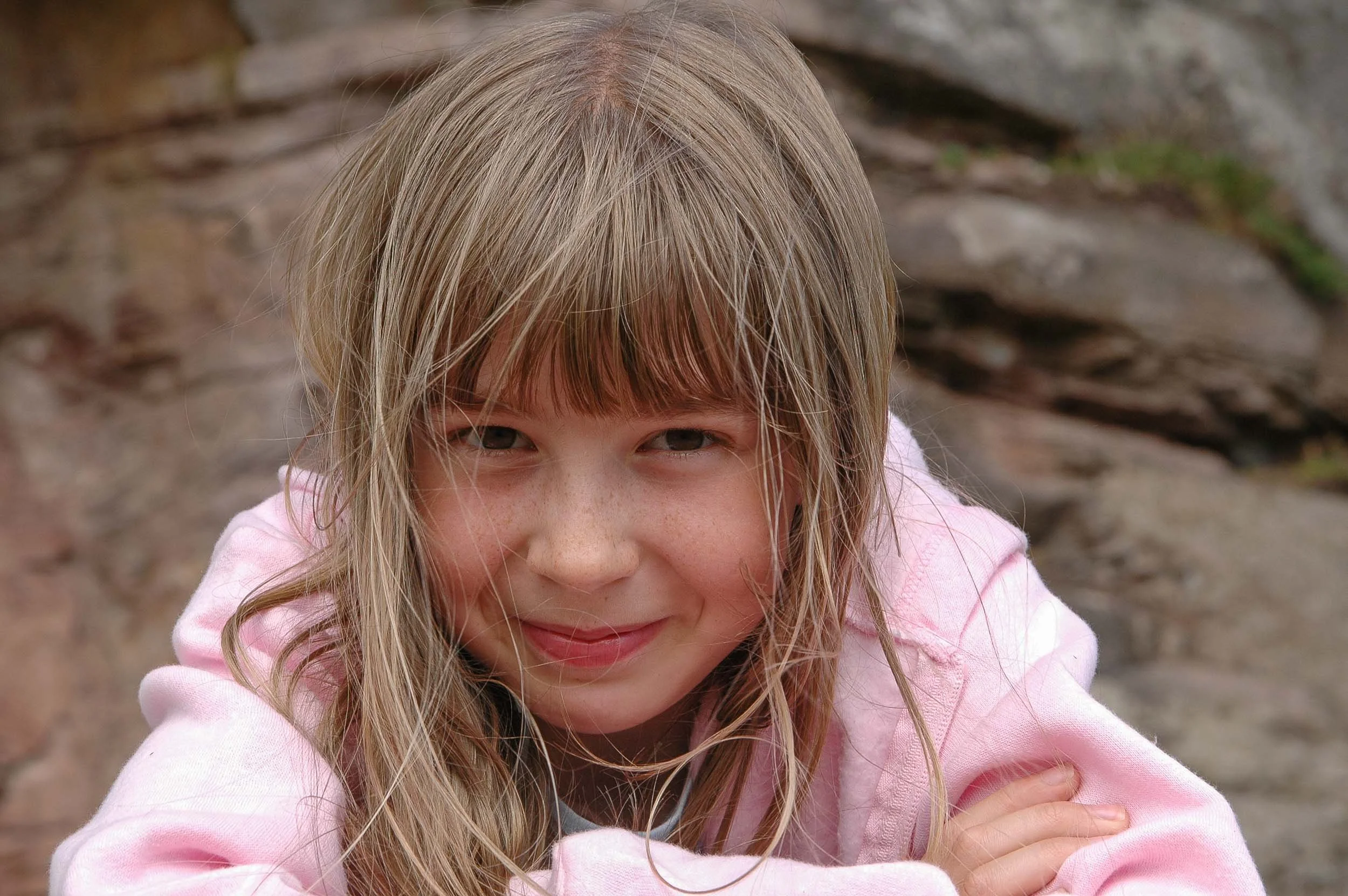 A young girl with blonde hair and freckles, wearing a pink hoodie, smiling outdoors in front of a rocky background.