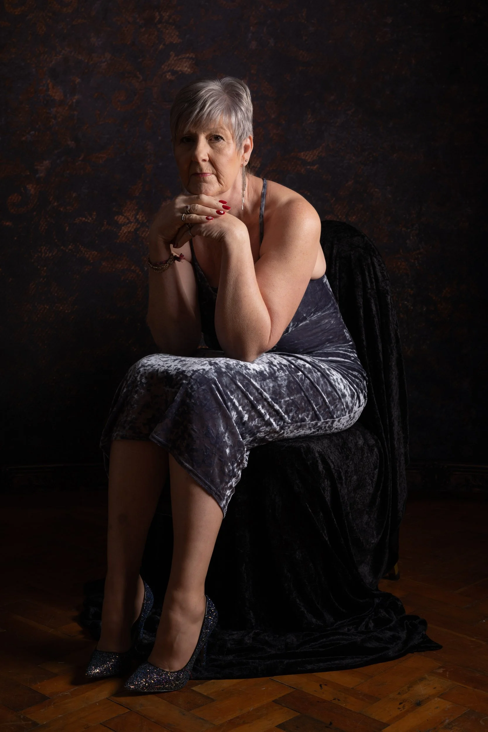 An elderly woman with short gray hair sitting on a black chair with a dark textured background. She is wearing a velvet dress with a crisscross strap, decorated with jewelry and sparkly high heels. She rests her chin on her hands, looking at the came