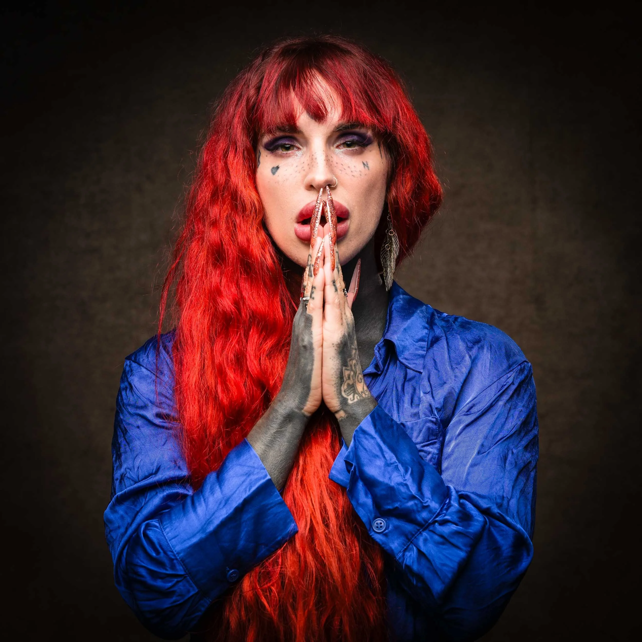 A woman with long red hair, tattoos, and face makeup, wearing a blue shirt, with her hands pressed together in front of her face in a prayer-like pose, against a dark background.