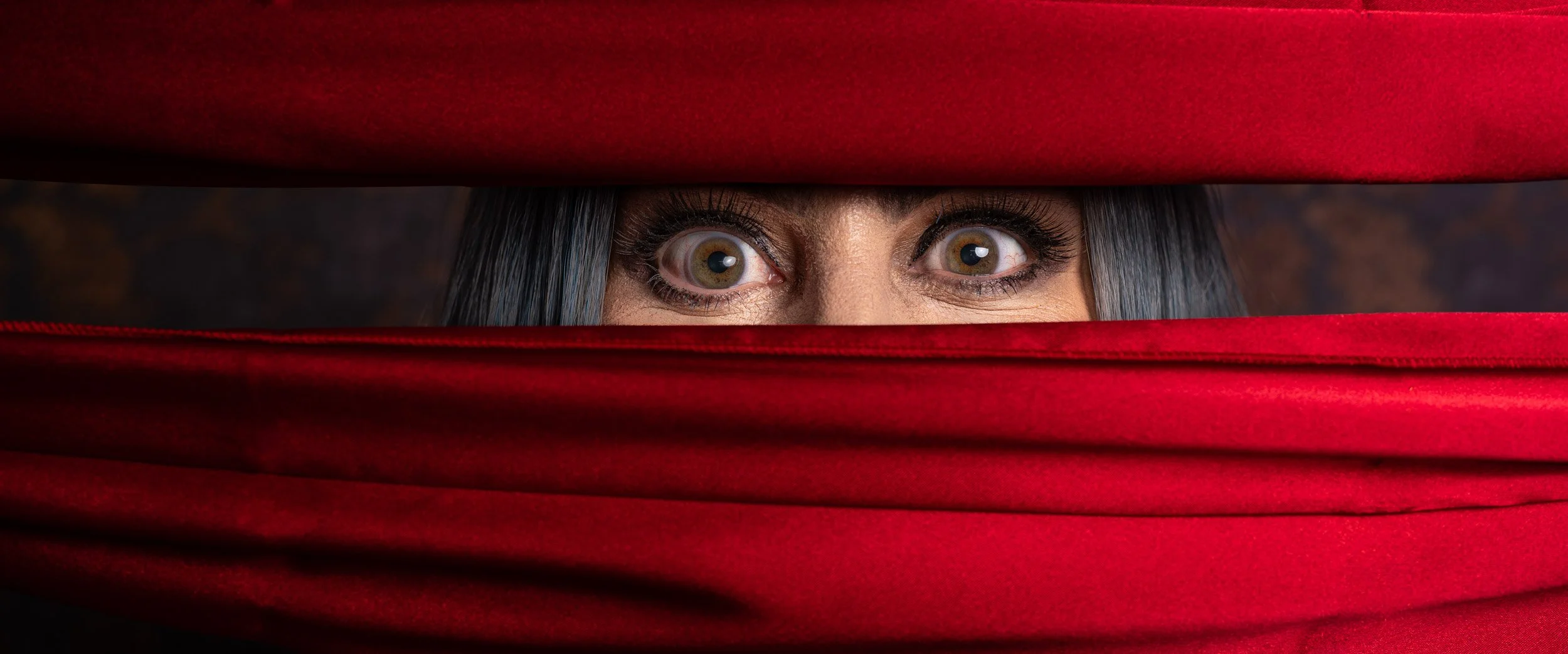 Person peeking through a slit in a red curtain, with wide eyes and dark hair.