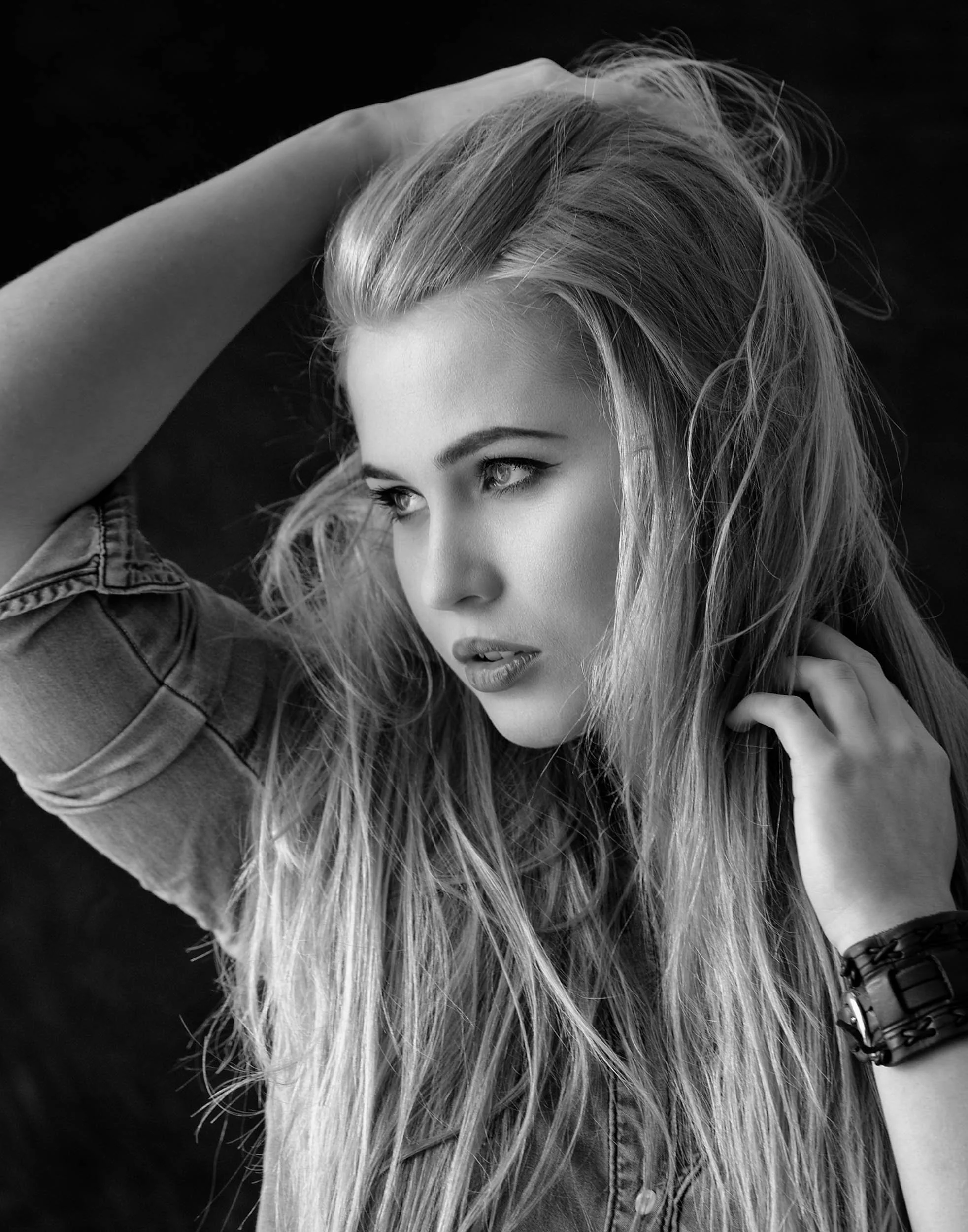 Black-and-white portrait of a young woman with long, wavy hair, with her left hand on her head and her right hand touching her neck, wearing a denim shirt and a black bracelet.